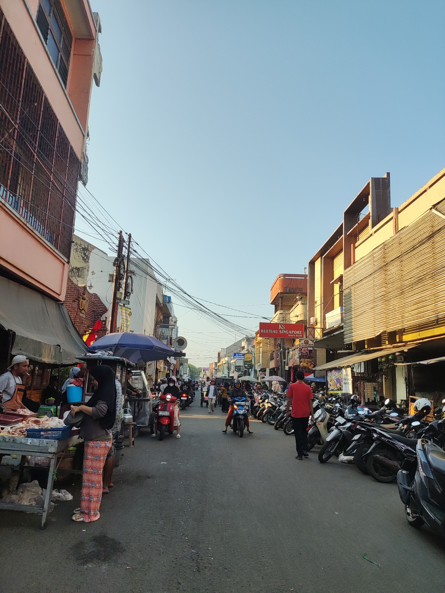 This is a street scene in the city of Cirebon, Indonesia, likely a market area. The image is dominated by a road filled with parked motorcycles and some moving traffic, primarily scooters. On the left side of the road, a vendor is seen at a stall with raw meat displayed. Several people are interacting with the vendor and the stall. A woman in a hijab is standing near the stall, holding a plate. Further down the street, more stalls and buildings line both sides. Signs are visible, one of which reads "KUETIAU SINGAPORE KOPI JAM". The buildings appear to be a mix of commercial establishments, with some showing signs of age and wear. The sky is clear and blue, suggesting it is daytime, likely late afternoon given the angle of the sunlight casting shadows. The overall atmosphere is that of a bustling urban environment.