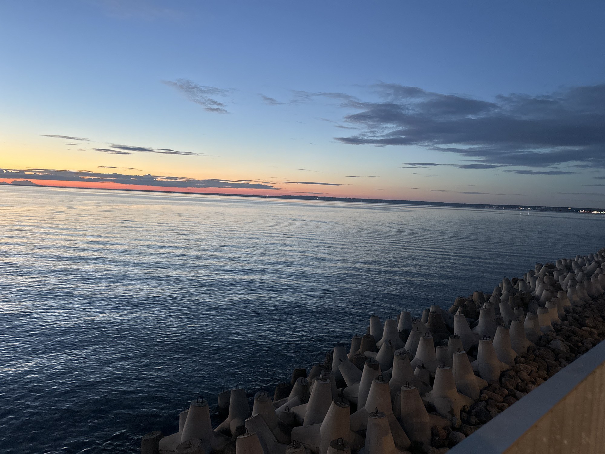 The image captures a serene sunset over the Baltic Sea, viewed from a breakwater constructed with concrete tetrapods. The sky displays a gradient of colors, transitioning from a soft orange and pink horizon where the sun has recently set, to a deep blue overhead. Wispy clouds are scattered across the sky. The sea is calm, reflecting the sky's colors. The tetrapods, a common type of coastal defense structure, are arranged in rows along the right side of the frame, forming a protective barrier. Beyond the sea, a distant coastline is visible with faint lights indicating a city or settlement, which is consistent with the location in Tallinn, Estonia, particularly around the Tallinn Bay area where such breakwaters are found. There are no people or active subjects depicted in the scene. The environment is natural and engineered, showcasing a coastal landscape at twilight.