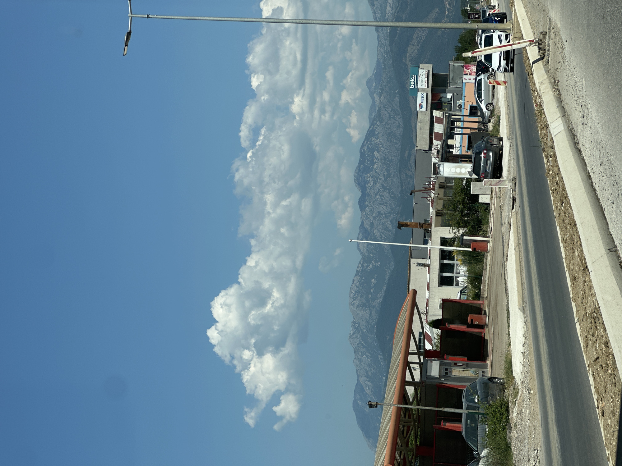 The image depicts a street scene in Bratešići, Montenegro, on a clear day with a bright blue sky and scattered white clouds. In the foreground, a paved road is visible, bordered by a construction zone with loose gravel and concrete barriers. Several cars are parked or driving on the road. To the left of the road, there is a car wash facility with an orange-topped structure. Behind the car wash, a low-rise building with a light-colored facade and windows is visible. Further back, a larger building with signage that includes "beko" and "BESIX SOLE CASA" is present. In the background, a mountain range with rugged peaks stretches across the horizon. A tall street lamp stands on the right side of the image. The overall scene suggests a commercial or industrial area on the outskirts of a town, with a backdrop of natural mountainous terrain.