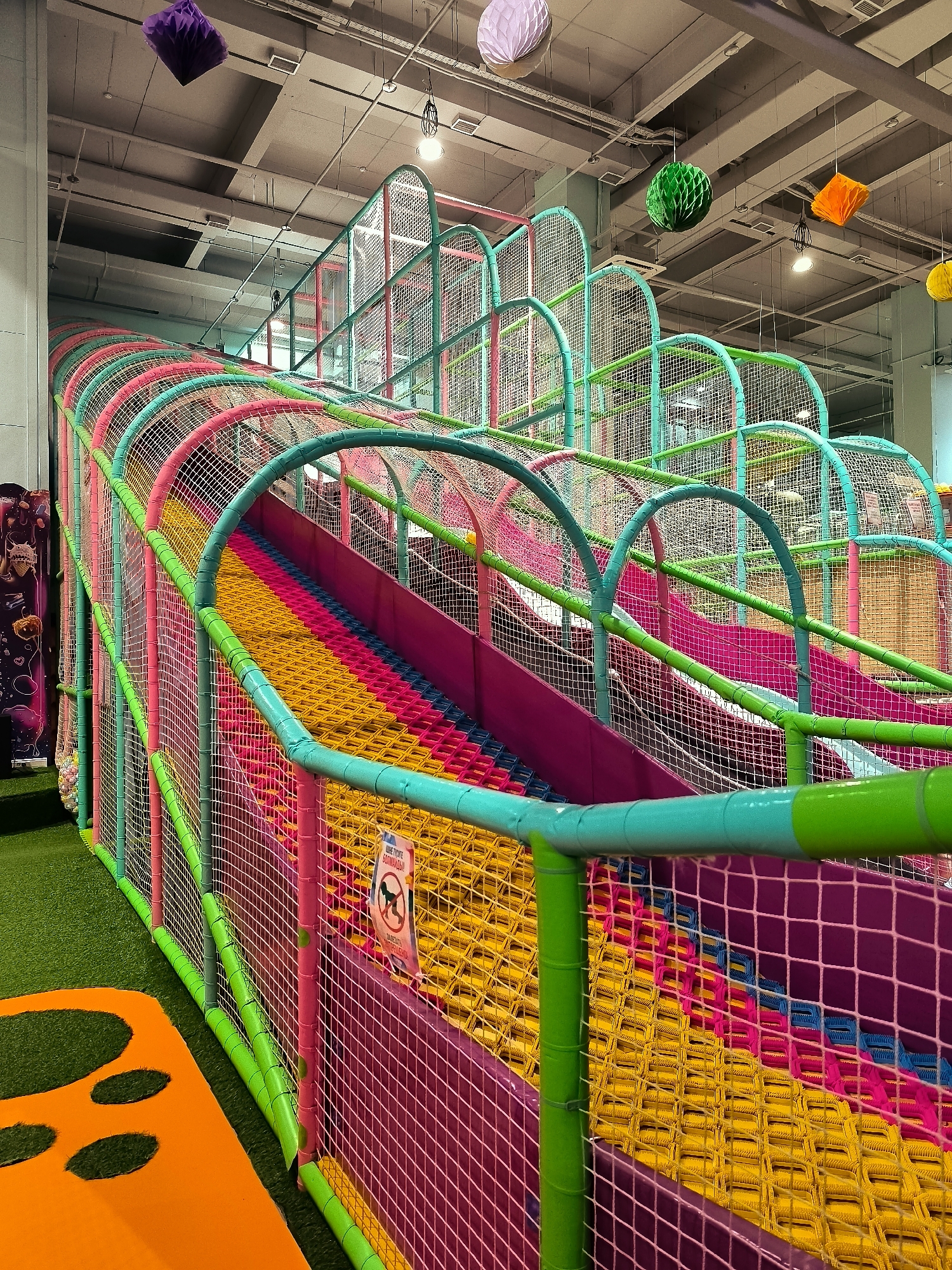 The image depicts a colorful indoor playground slide structure. The slide is enclosed by netting and supported by brightly colored poles in pink, teal, and lime green. The slide surface itself is a vibrant rainbow of yellow, pink, and blue sections. Above the structure, decorative paper balls in purple, green, and orange are suspended from the ceiling, along with lighting fixtures and ventilation systems. To the left, a patch of artificial green turf is visible, along with an orange play mat with cutouts. A sign with a dog symbol crossed out and text in Russian is affixed to the netting on the right side of the slide. The overall impression is of a well-lit, child-friendly play area.