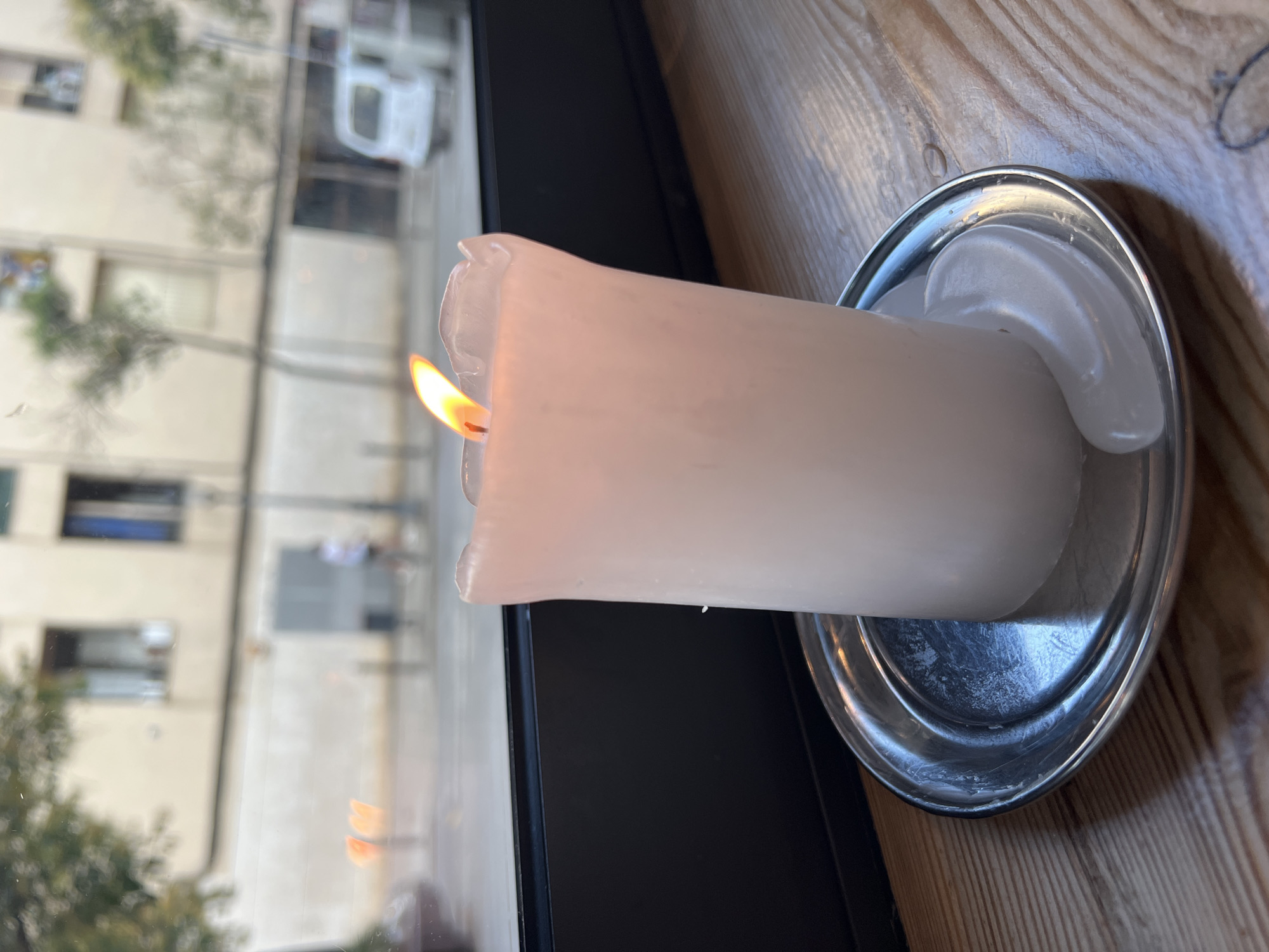 A lit white candle in a silver holder sits on a wooden table, in front of a window with a view of a street in Barcelona, Spain. The candle's flame is bright orange and yellow, and wax has melted down the sides of the candle, forming drips. Behind the candle, the window shows a blurred street scene with buildings, trees, and vehicles. The buildings are light-colored, with several windows visible. A white van is parked on the street. The overall lighting suggests it is daytime.