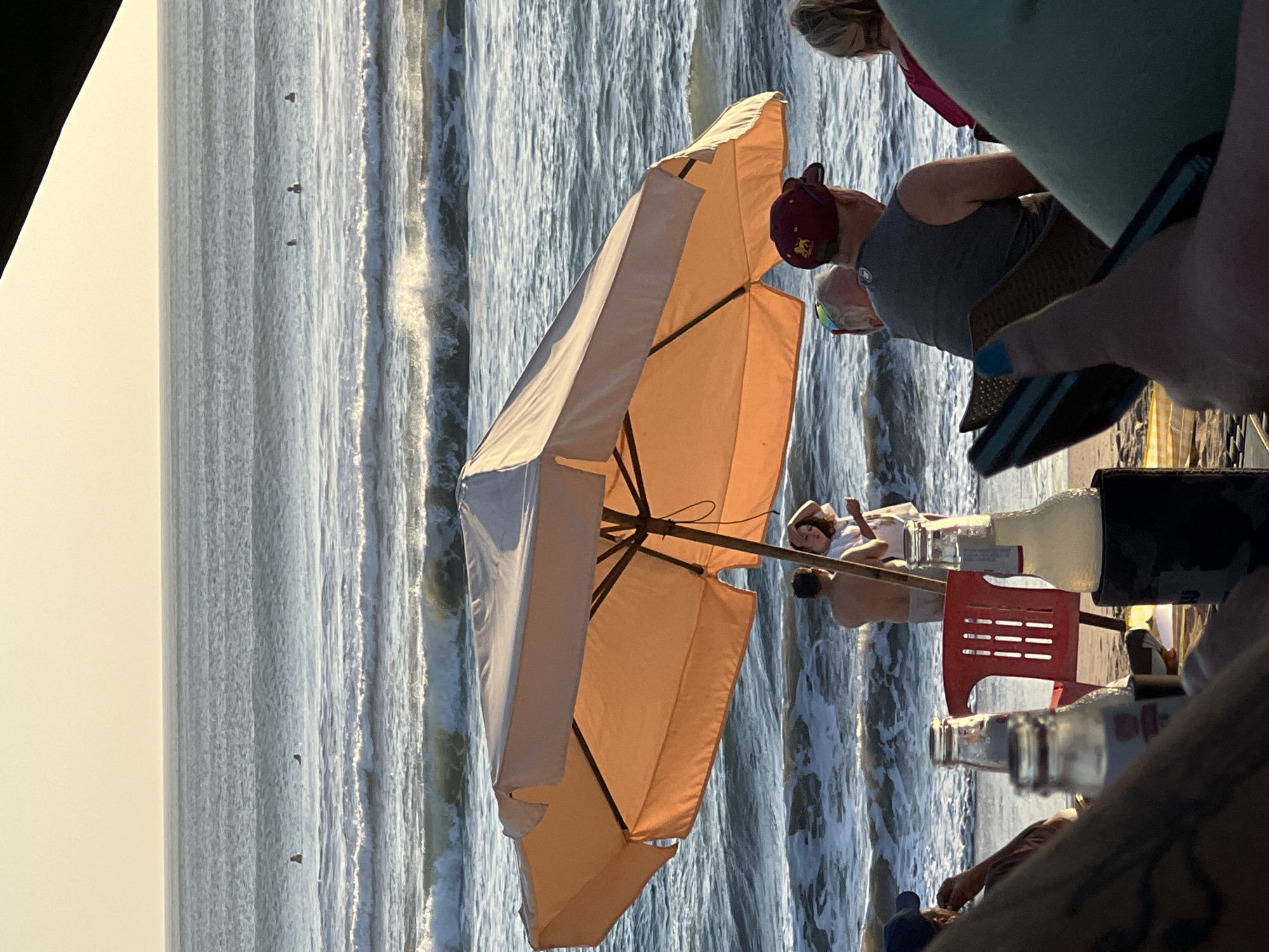 A group of people are gathered at a beachside location, possibly a cafe or restaurant, in Kuta, Indonesia. The scene is set at sunset, with the sky transitioning from a pale yellow to a light orange. The ocean is visible in the background, with gentle waves rolling towards the shore.
In the foreground, several people are seated, their backs to the camera, enjoying the view. A hand with blue nail polish is prominently positioned, giving a thumbs-up gesture. On a table, two glass bottles, one clear and one with a yellowish liquid, are placed.
Further back, under a large, peach-colored umbrella, two individuals are standing near the water. One appears to be a shirtless man, while the other, a woman in a white outfit, is looking at him. A red plastic chair is also visible. In the middle ground of the ocean, several surfers can be seen in the distance, enjoying the waves. The overall atmosphere suggests a relaxed and enjoyable evening by the sea.
