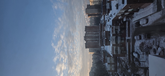A winter cityscape under a blue sky streaked with clouds. Tall apartment buildings are juxtaposed with smaller residential structures, all dusted with snow. Cars and streets are also partially covered. The scene suggests a cold, early morning or late afternoon with a soft, diffused light. The perspective indicates the photograph was taken from a high vantage point.
