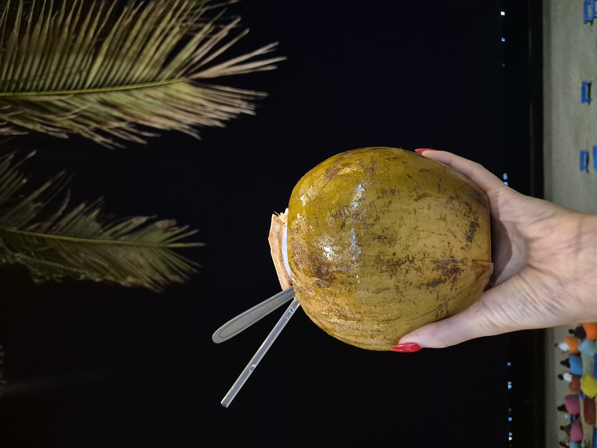 A person's hand with red nail polish holds a green coconut with two straws and a metal spoon inserted into its opening. The coconut is positioned in the foreground, with palm fronds visible against a dark night sky. In the background, a beach scene unfolds with blurred lights of a city or town along the coastline, and several beanbag chairs are scattered on the sand. This scene is likely on a beach in Đà Nẵng, Vietnam, given the presence of palm trees, the beach setting, and the proximity to a populated coastal area.