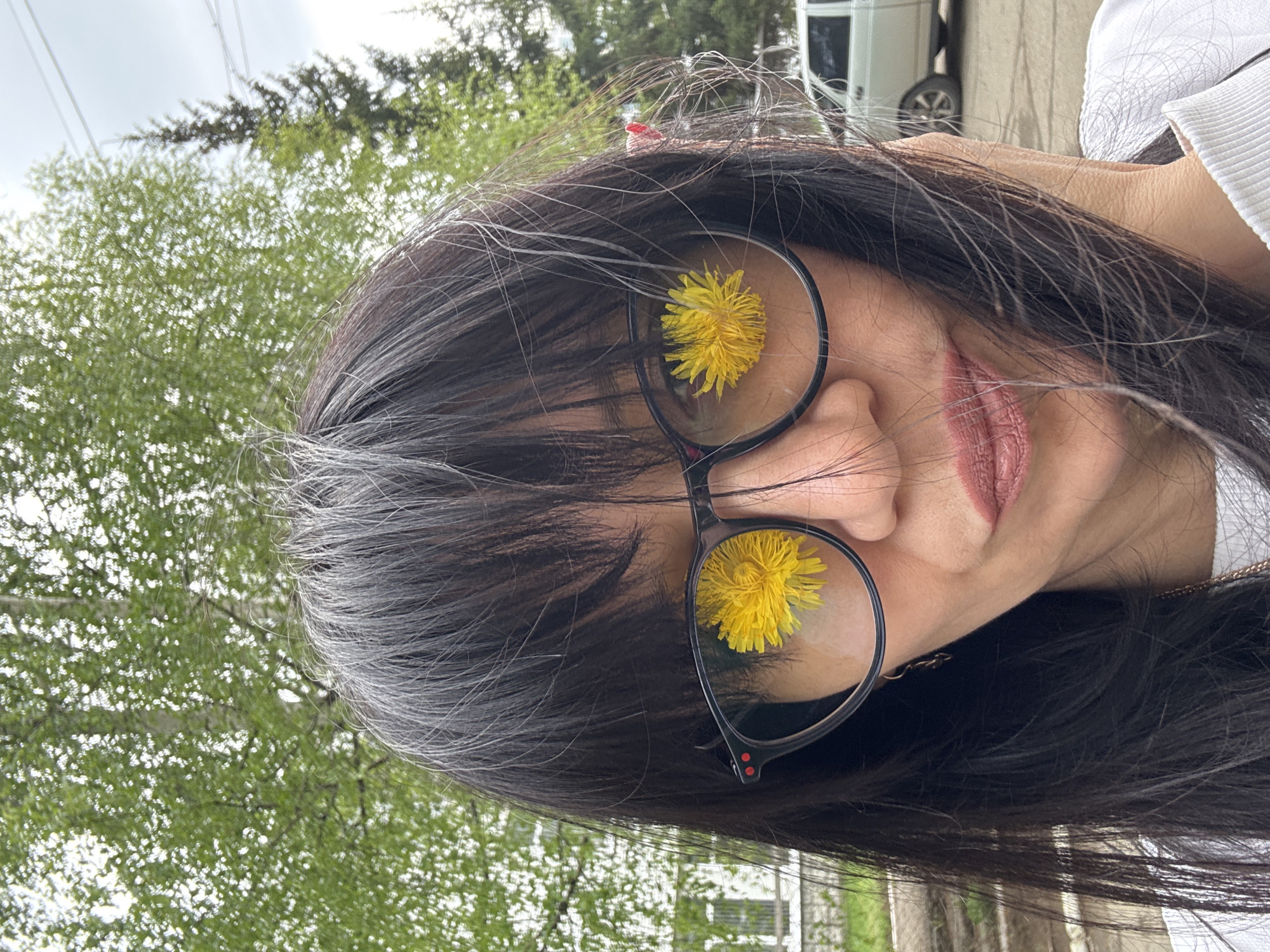 A headshot of a woman with dark hair and glasses with a dandelion in each lens, taken outdoors in the city of Chaikovsky, Russia. The background shows trees and a white van. The woman is wearing a white shirt.