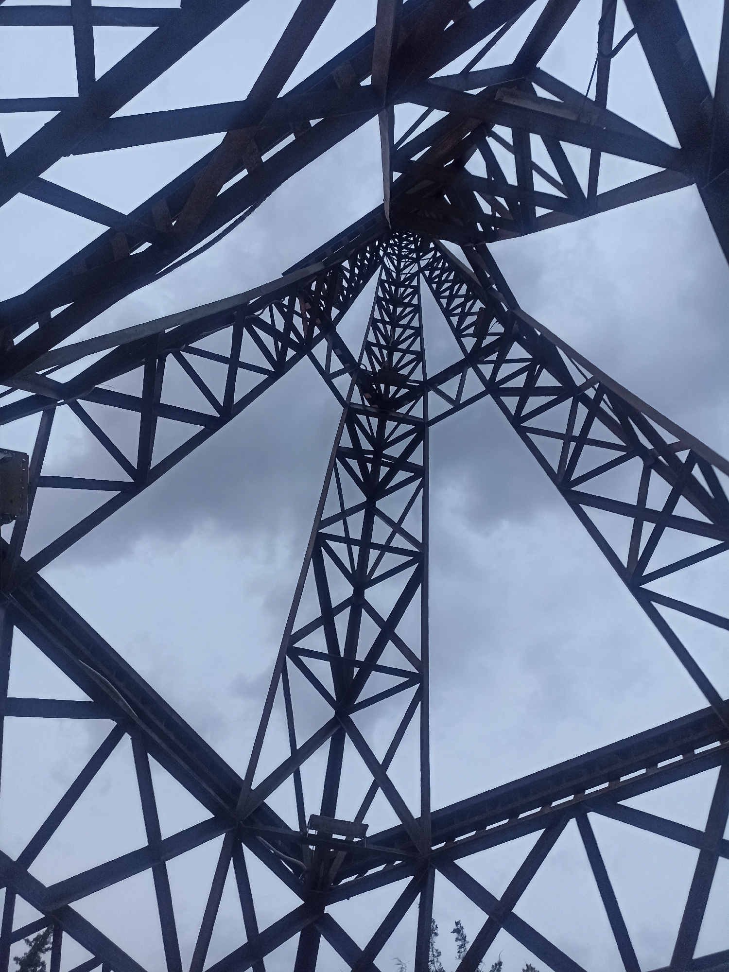 The image is an upward-looking, low-angle shot of a large, dark metal lattice tower against a cloudy sky. The tower is constructed from numerous intersecting beams, forming a complex geometric pattern that recedes towards the top. The sky is overcast, with grey clouds dominating the background, creating a somber and industrial atmosphere. The perspective emphasizes the scale and height of the tower, with the sky appearing as a void between its structural elements. There are no people visible in the image. The scene appears to be outdoors, possibly in an industrial or infrastructure area. Given the provided context that the image is located in Osh City, Kyrgyzstan, this structure could be a telecommunications tower, a power line pylon, or part of another large-scale industrial installation typical of such urban environments.