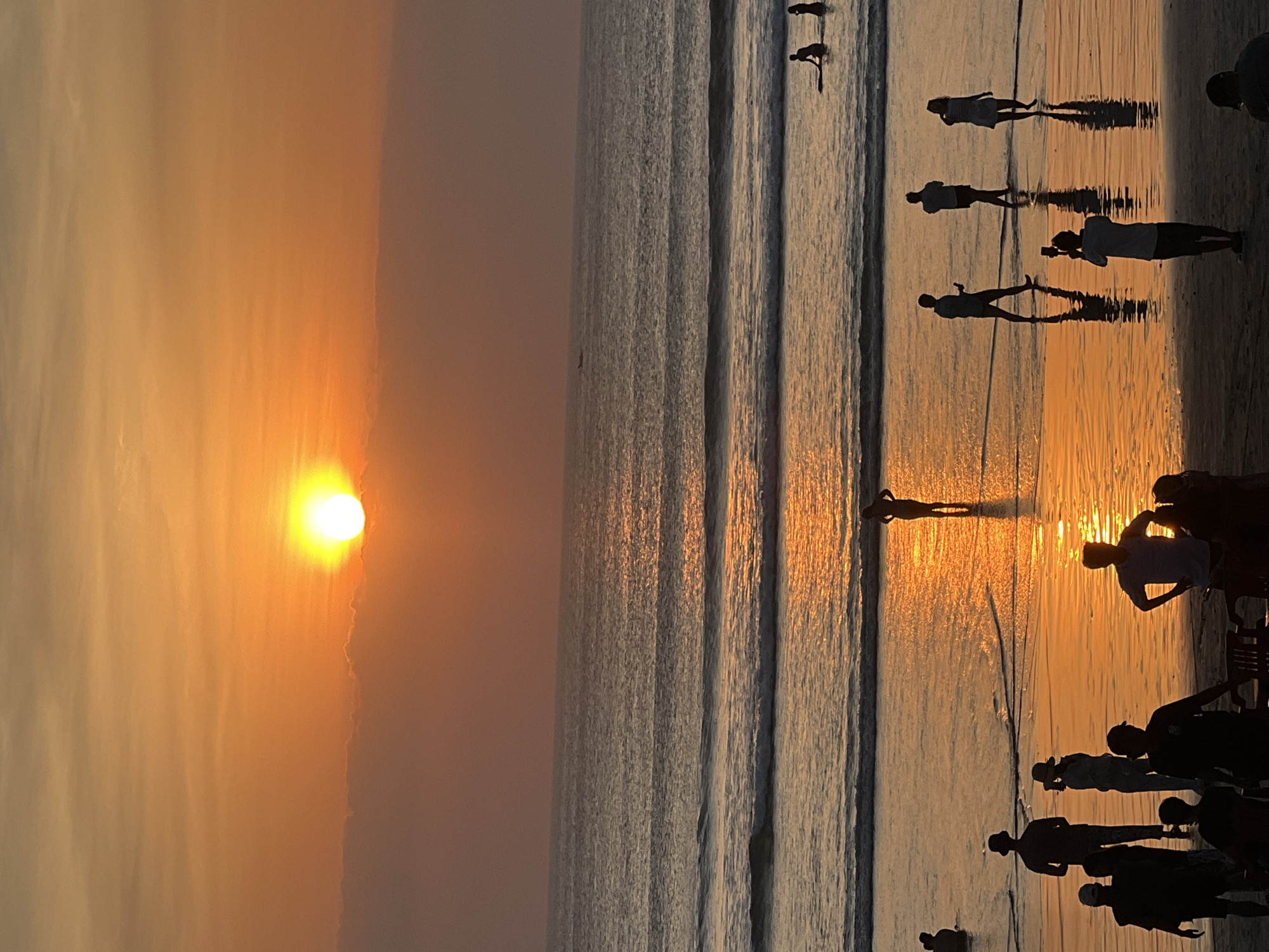 At sunset on Kuta Beach, Bali, Indonesia, the sky is painted in warm hues of orange and yellow, with the sun a bright disc partially obscured by clouds. The ocean, reflecting the vibrant sky, features gentle waves breaking on the shore. Silhouettes of people are visible both on the wet sand and in the shallow water, enjoying the end of the day. Some stand alone, others in small groups, their figures elongated by the low angle of the sun. A few individuals are wading in the surf, while others are closer to the shoreline. In the distance, a small boat is visible on the water. The scene is one of peaceful enjoyment of the natural beauty of the beach.