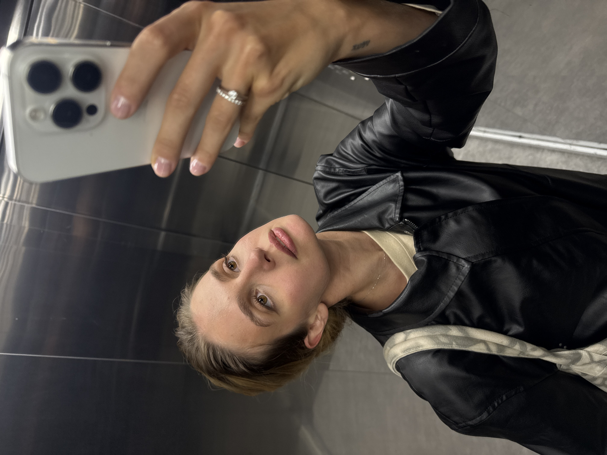A fair-skinned person with blond hair is taking a selfie in an elevator. The person is wearing a black leather jacket over a light-colored top. Their left hand is holding a white smartphone with multiple camera lenses. The person is wearing a ring on their left ring finger and there's a tattoo visible on the left wrist. The elevator has brushed metal walls and a horizontal rail in the background.