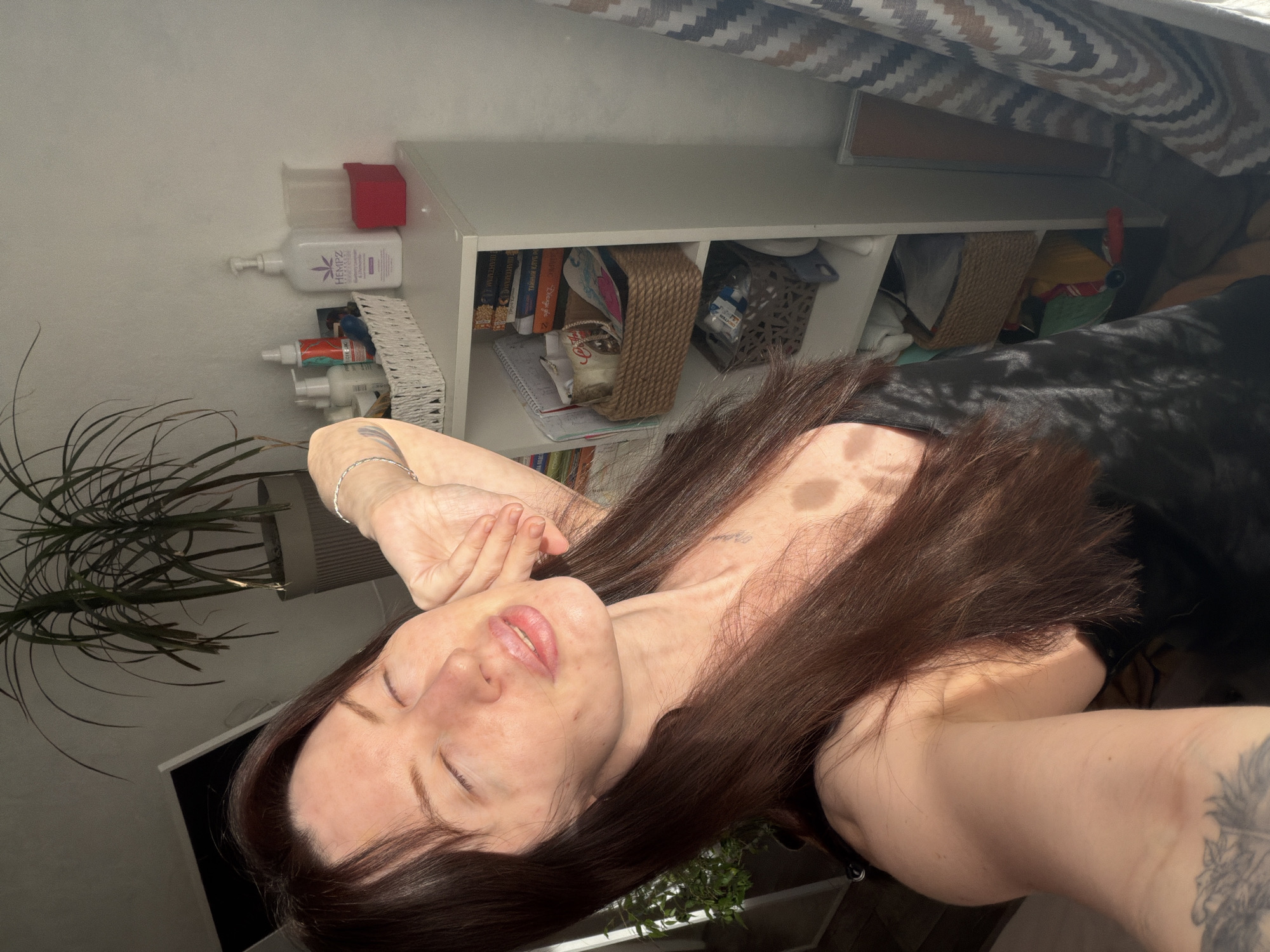 The image is a selfie-style photo of a woman indoors. She is lying down, facing the camera with her eyes closed. She has long, dark brown hair and fair skin. Her left hand is near her face. She is wearing a dark garment. To the left of the woman is a potted plant. Behind her is a white shelving unit containing books, baskets, and bottles. The location cannot be determined as Perm, Russia without additional information.