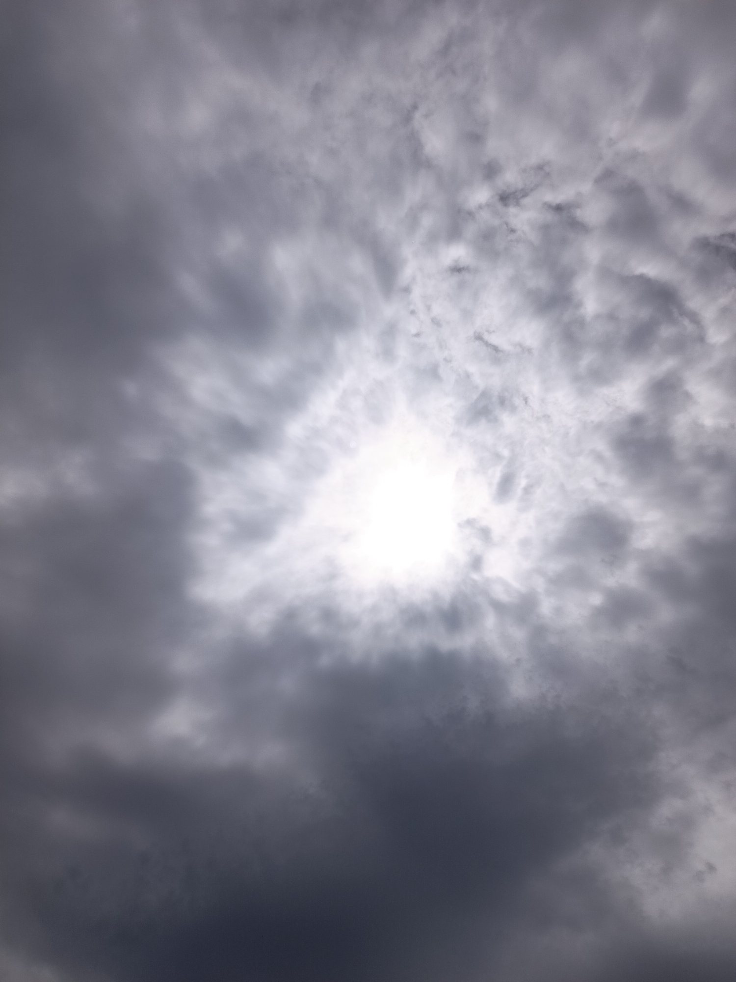 This media file shows a view of the sky from below, looking upwards. The sky is filled with a mix of dark, heavy clouds and lighter, more scattered clouds, creating a dramatic and somewhat overcast atmosphere. In the center of the image, a bright, luminous patch of sky breaks through the clouds, suggesting the sun is present, though its direct rays are diffused by the cloud cover. This creates a halo-like effect around the brightest point. The clouds are varied in texture and density, with some appearing wispy and others thick and imposing. The overall impression is of a dynamic sky, possibly transitioning between weather conditions. There is no visible text. The scene is an outdoor, natural setting, focusing solely on the sky. The lighting suggests it is daytime.