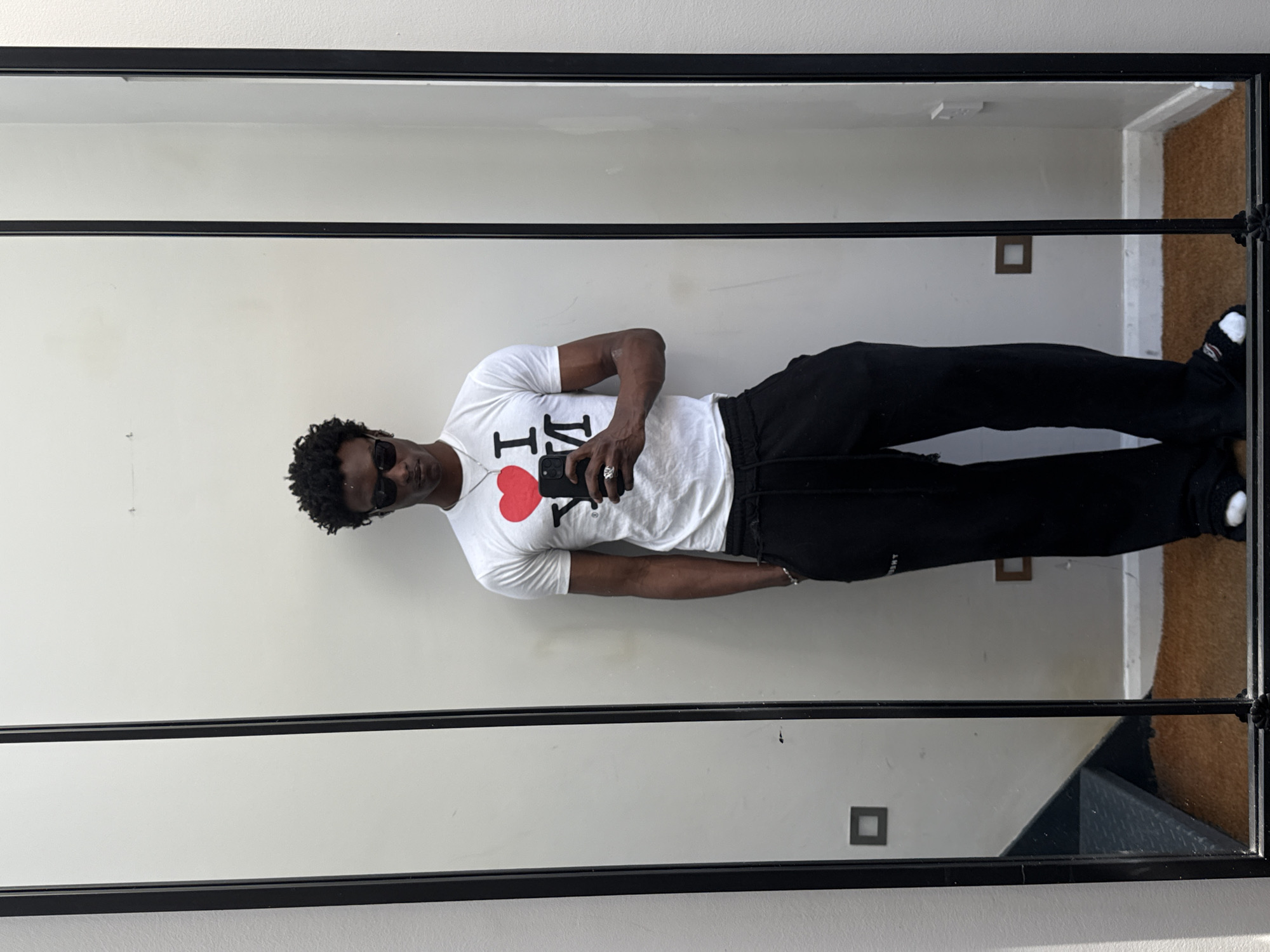 A man stands in front of a mirror taking a photo of himself. He wears sunglasses, a white t-shirt with an "I love NY" print, black sweatpants, and socks with sandals. He holds a phone in his right hand. The background includes a plain white wall, a doorway leading to a room with cork flooring, and a dark stairwell visible in the mirror's reflection. The room is possibly located in London, United Kingdom.