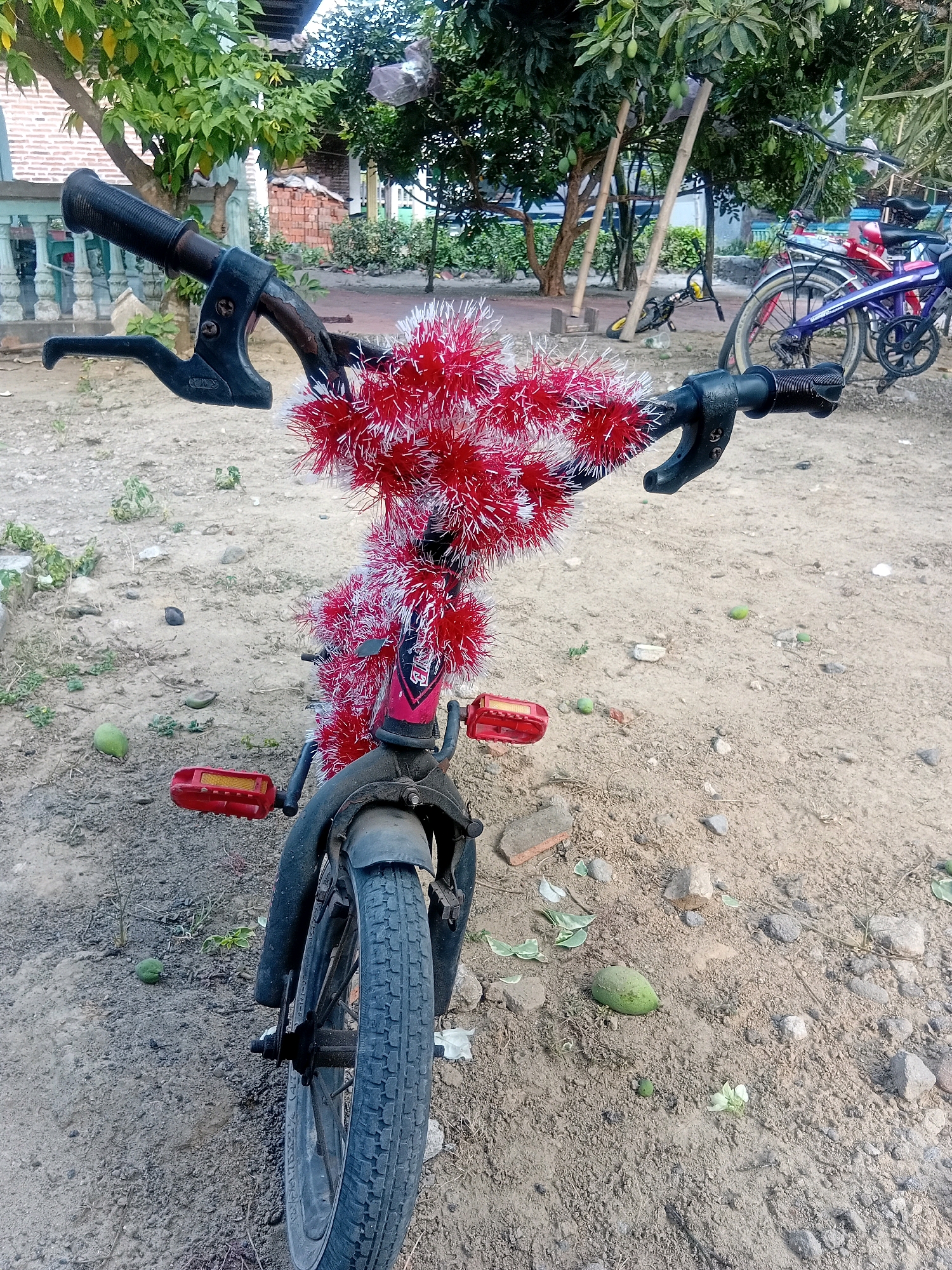 A red and white tinsel-decorated children's bicycle is parked on a dirt ground in Banjarwungu, Indonesia. The bicycle has black handlebars with red streamers attached. The front wheel and tire are visible, along with red pedals. In the background, there are trees, buildings, and other bicycles. The ground is covered in dirt, small rocks, and some fallen leaves and green fruits.