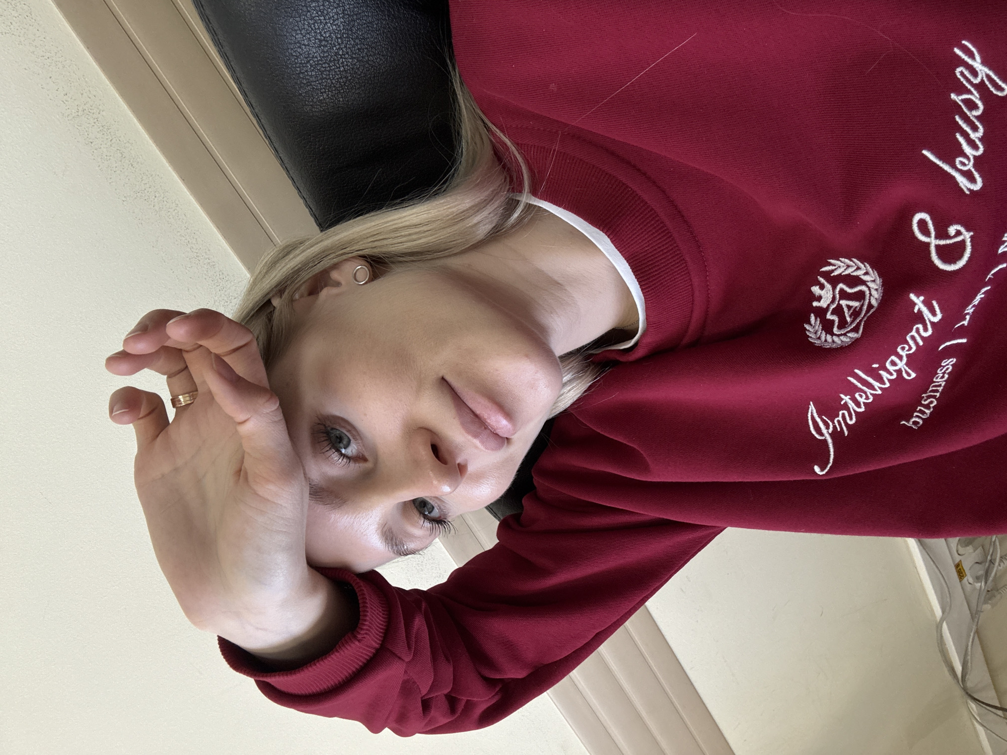 A blonde woman with fair skin is facing the camera, her face angled. Her right hand is raised near her face. She has light eyes, and her hair is light blonde. She is wearing a red sweatshirt with white lettering and a design on it. She is sitting in a black leather chair. The location is not discernible from the image. The environment is generic and does not provide location information.