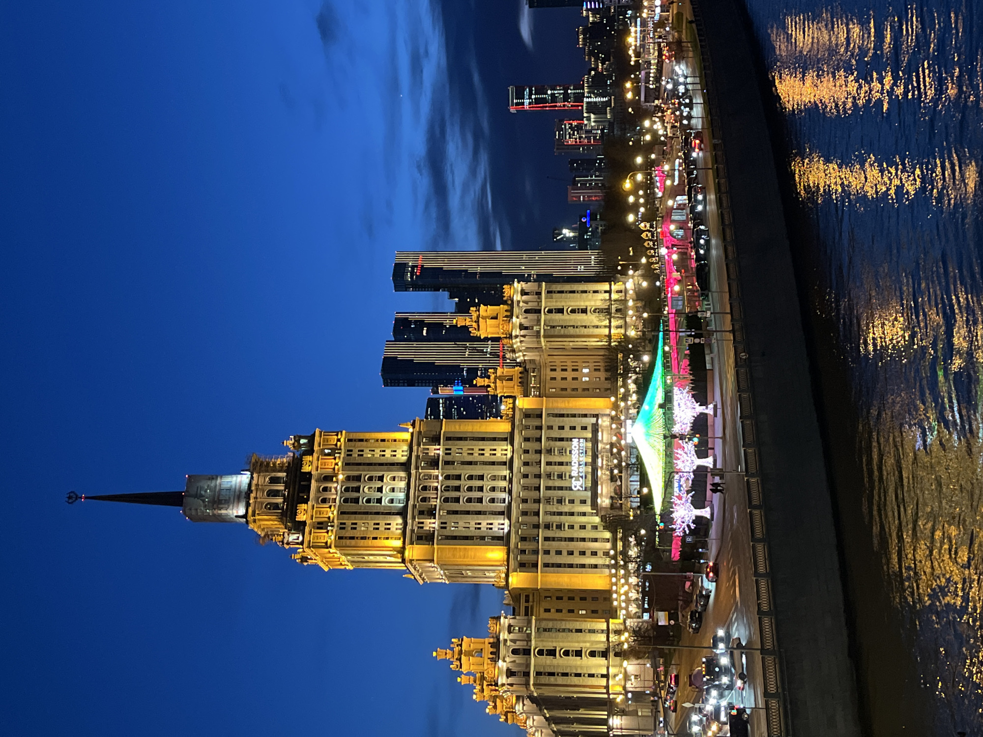 The image is a night view of a lit-up cityscape in Moscow, Russia. A prominent Stalinist skyscraper with a spire dominates the left side of the frame. The building is brightly illuminated in yellow. Behind it, modern skyscrapers rise into the dark blue sky with scattered clouds. To the right of the building, a brightly lit street runs alongside the water, with festive decorations, including illuminated trees. The water is reflecting the lights of the city. The scene is calm and urban.