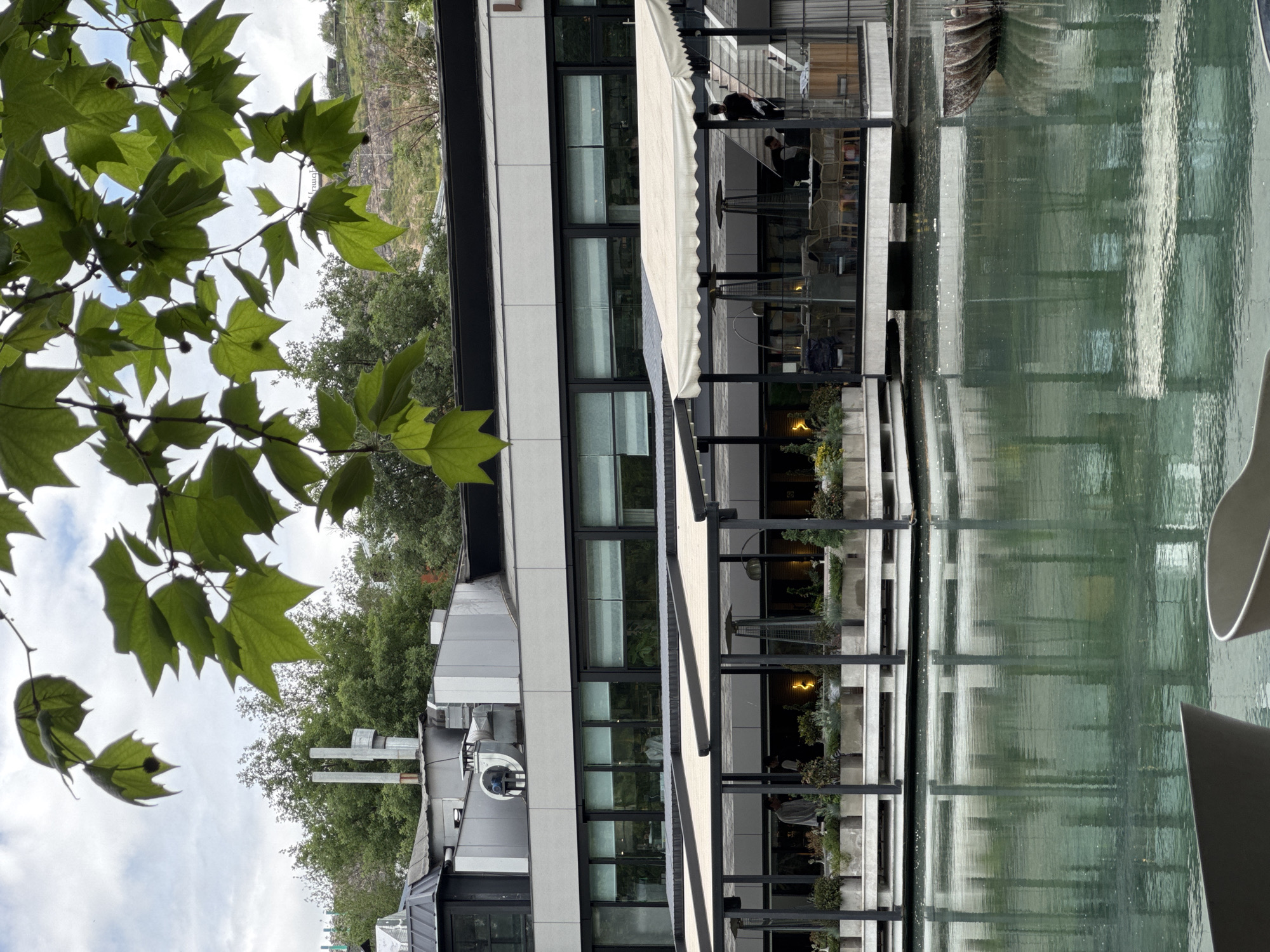 The image depicts a building with a sleek, modern architectural style in Yerevan, Armenia. The building features multiple floors with large windows and a retractable awning covering an outdoor seating area. The outdoor space is furnished with tables and chairs, where several people can be seen seated. The building is situated next to a body of water, possibly a canal or a decorative pool, reflecting the building's facade. The foreground includes the lush green leaves of a tree. The sky is partly cloudy, indicating the weather is mild.