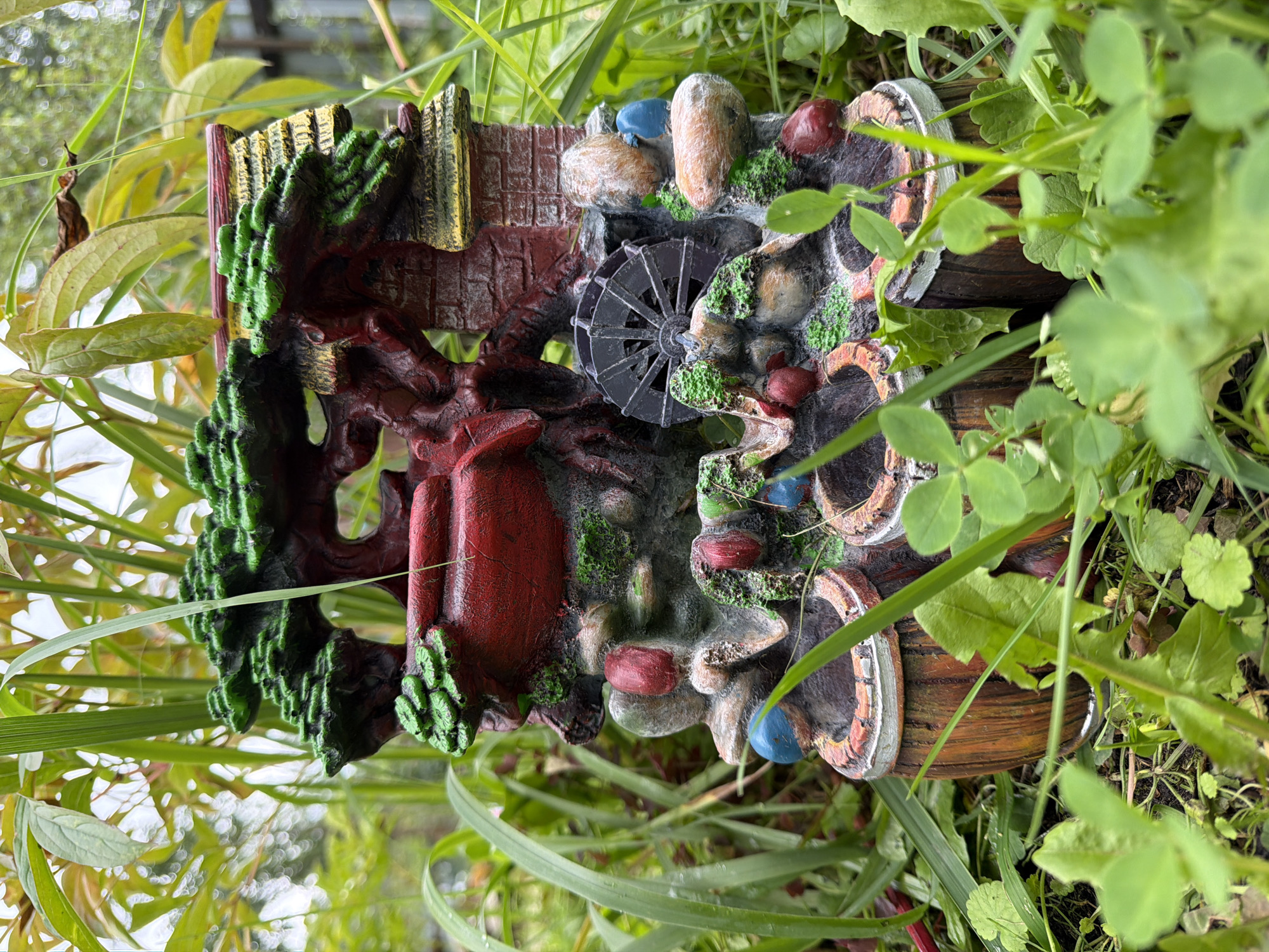 A decorative garden ornament shaped like a miniature water mill is situated in a grassy area surrounded by greenery. The ornament depicts a small building with a thatched roof, a water wheel, and several red, blue, and brown decorative elements resembling rocks or fruits. The ornament is brightly colored with green for foliage, red for the building and fruit-like objects, and brown for wooden barrels at the base. The background consists of various green plants and foliage, with hints of a fence and possibly trees in the distance. The image appears to be taken outdoors, likely in a garden or park setting. There are no people or animals visible in the image. The lighting suggests it is daytime, possibly overcast due to the soft, diffused light. The ornament is the primary subject, placed centrally in the frame. The location in the city of Pokrov, Russia, is not evident from the visual details of the image itself.