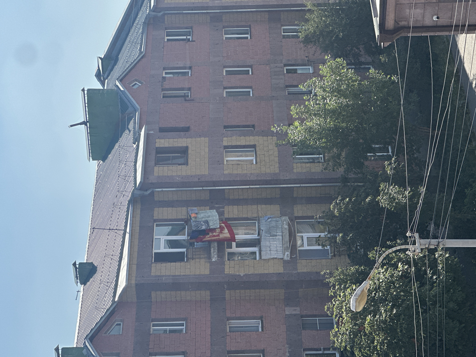 The image depicts a multi-story apartment building with a facade of alternating pink and yellow brickwork, situated in the city of Gyumri, Armenia. The building has a dark-colored, sloped roof with several green structures atop it, one of which appears to be a chimney. A balcony on one of the upper floors displays several pieces of laundry, including a large red tapestry and a lighter colored fabric, hanging to dry. Trees with lush green foliage partially obscure the lower levels of the building. In the foreground, a street lamp with a curved pole stands beside a utility pole supporting multiple electrical wires. The sky is clear and blue.