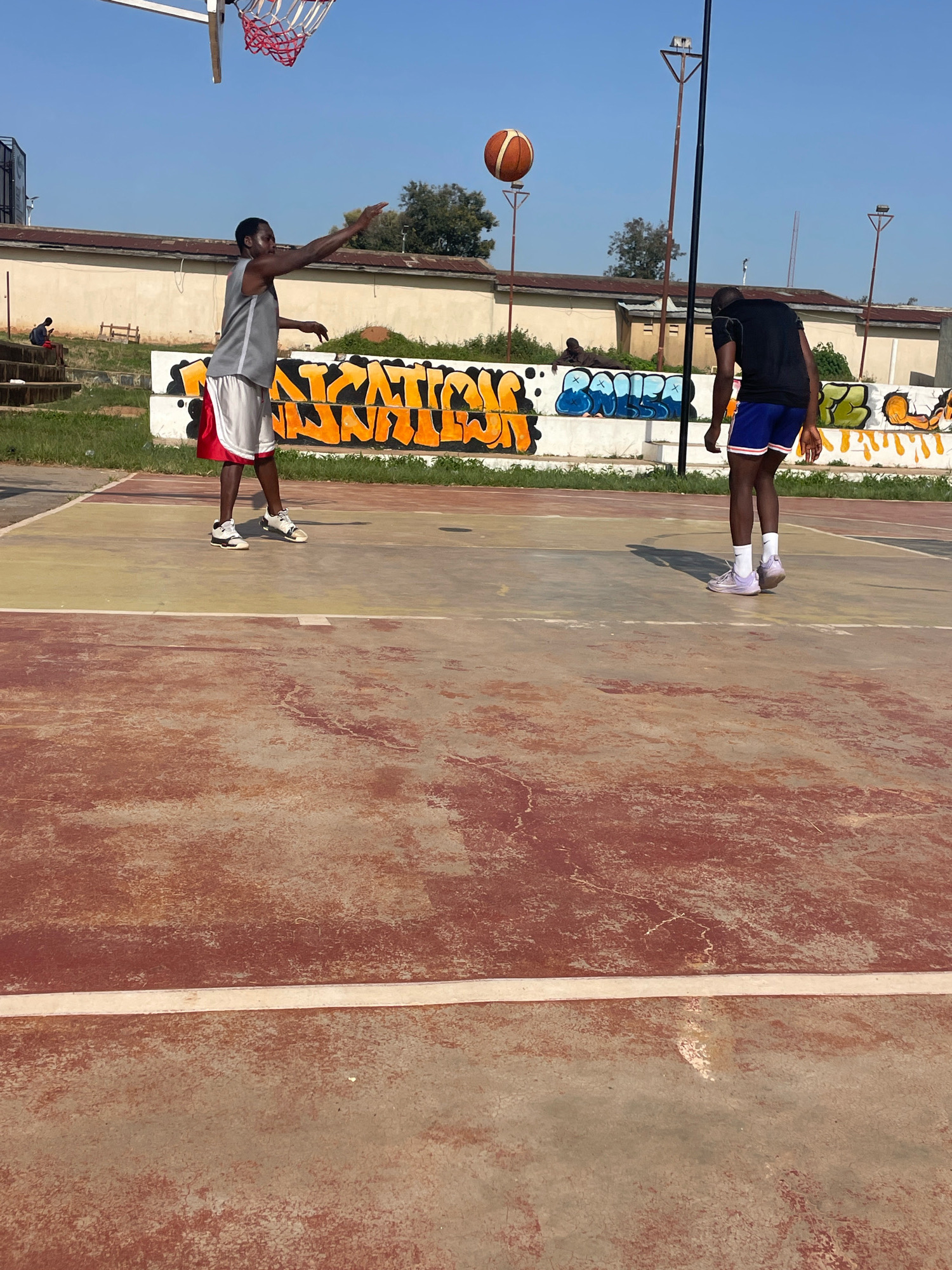 The image depicts an outdoor basketball court with two young men playing. The court surface is worn and shows patches of red and beige concrete. The background features a low, beige building with a tiled roof and graffiti-covered walls, some with vibrant orange and blue lettering. A basketball hoop with a red net is visible on the left.
One young man, wearing a grey tank top and red and white basketball shorts, is in the process of shooting a basketball. He is positioned on the left side of the court, mid-action with his arm extended upwards towards the hoop. He wears white and black basketball shoes.
Another young man, wearing a black t-shirt and blue basketball shorts with white trim, stands on the right side of the court, facing away from the camera, observing the shot. He wears white socks and light purple athletic shoes. A basketball is suspended in the air above and to the right of the first player, indicating the shot has been released.
The sky is clear and blue, suggesting a sunny day. The general environment appears to be an urban or semi-urban setting, potentially part of a community park or recreational area in Jos, Nigeria. The presence of graffiti and the worn state of the court suggest a well-used, possibly older facility.