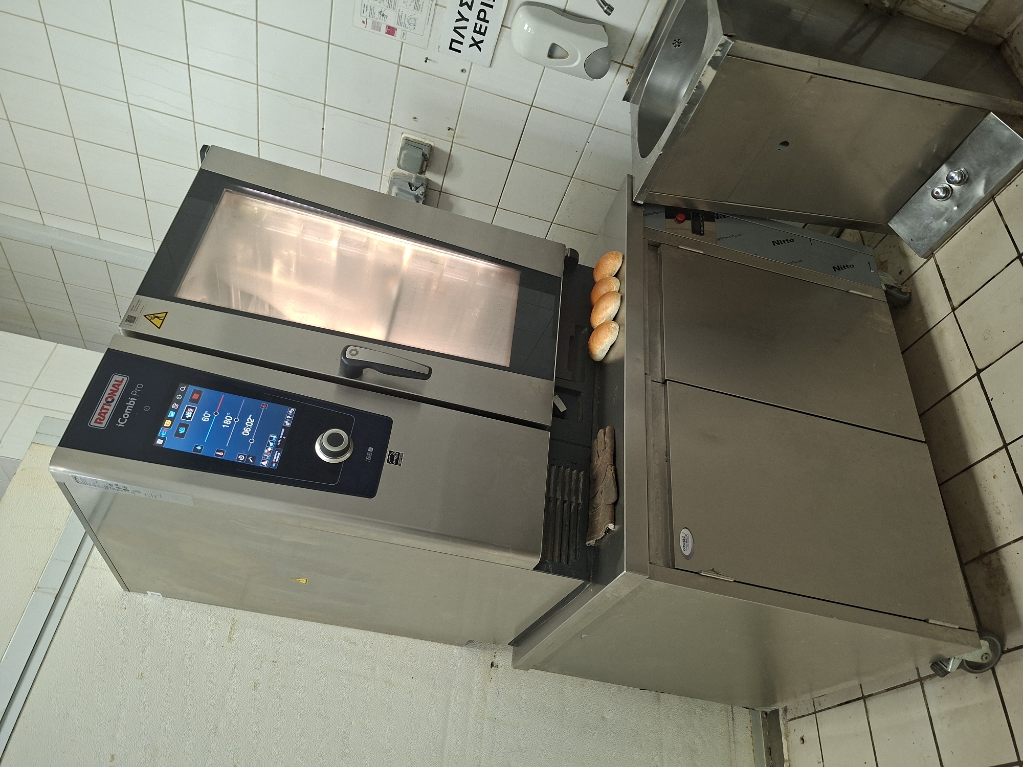 The image depicts a professional kitchen setting, likely in Nicosia, Cyprus, featuring a Rational iCombi Pro combi oven prominently placed on a stainless steel cabinet. The oven has a digital display showing cooking settings such as 60°C and 180°, and a timer set to 06:02. Resting on the stainless steel surface in front of the oven are four golden-brown bread rolls. To the right of the oven, a stainless steel sink is visible, accompanied by a white soap dispenser mounted on a tiled wall. The wall behind the oven and to the left is tiled in a light color, while the floor is also tiled with a grid pattern. A pair of brown leather gloves is placed on the stainless steel cabinet to the left of the bread rolls. The overall environment appears utilitarian and functional, consistent with a commercial kitchen.