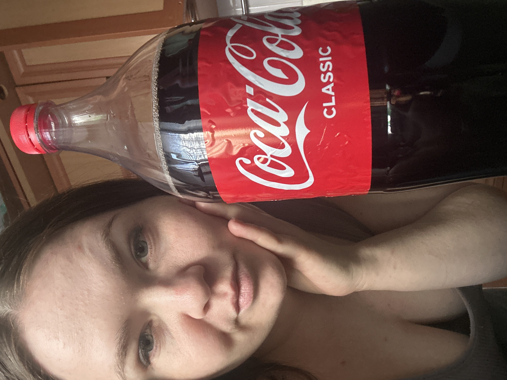 The image captures a rotated close-up view of a woman and a large Coca-Cola bottle indoors, set in Chaikovsky, Russia. A woman, visible from the chest up, looks directly at the camera with a neutral, slightly pensive expression, her left hand gently touching her cheek. Her dark hair frames her face.
A substantial, almost full, clear plastic bottle of Coca-Cola Classic is positioned prominently, seemingly resting on her shoulder or head, with its red screw cap visible on the left side of the frame. The bottle features the distinct red label with the white "Coca-Cola" cursive logo and the word "CLASSIC" printed in white uppercase letters beneath it.
The background is softly blurred, revealing light brown wooden elements, likely cabinetry or furniture, suggesting a domestic interior, possibly a kitchen or living space. The lighting appears to be natural, indicating it is daytime. No specific weather conditions are discernible.