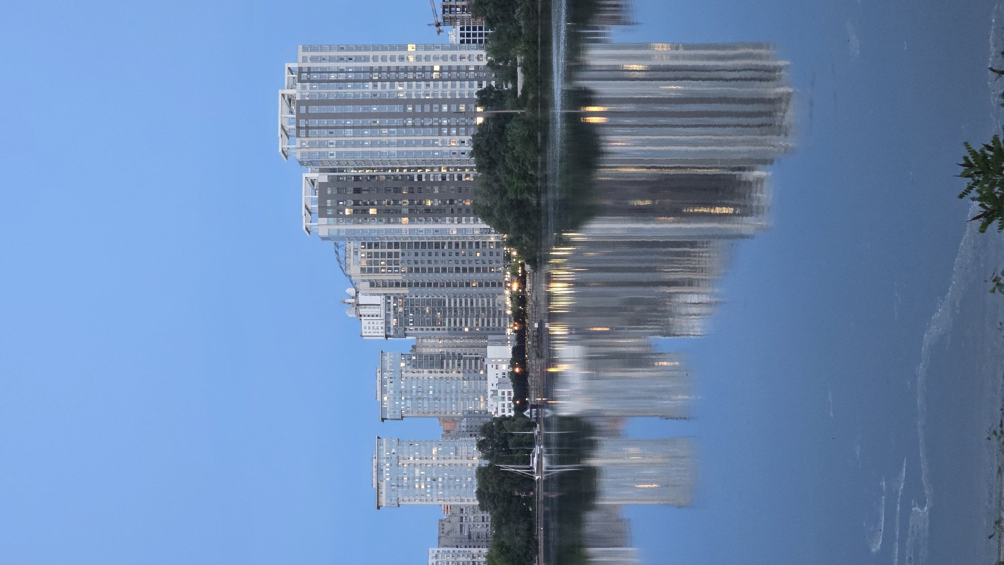 The image displays a modern urban skyline at twilight, reflected in a body of water. The upper portion of the frame is dominated by a clear, gradient blue sky, transitioning from a lighter shade at the horizon to a deeper blue overhead, devoid of clouds.
In the mid-ground, a dense cluster of high-rise residential or commercial buildings forms the urban profile. These structures exhibit contemporary architectural styles, predominantly featuring extensive glass facades, light-colored concrete or metal frameworks, and some darker grey sections. Numerous windows are illuminated with warm yellow light, indicating occupancy. Several prominent buildings feature distinctive white-framed roof structures. A partially visible construction crane on the far right indicates ongoing development. To the far left, two distinct, taller buildings with flat roofs are visible, interspersed with other structures.
Separating the buildings from the water is a narrow strip of dark green trees and dense foliage. Along this tree-lined bank, multiple artificial lights, consistent with streetlights or promenade lighting, emit a warm glow that reflects onto the water. A white sailboat with two masts is moored near the left bank, partially visible behind the foliage.
The lower half of the image is occupied by a dark blue body of water, which perfectly mirrors the skyline. The reflections of the buildings are prominent, slightly elongated, and diffused, with the illuminated windows appearing as shimmering vertical streaks of light. In the bottom left foreground, faint, lighter-colored circular patterns, indicative of surface foam or debris, are visible on the water. A small cluster of green foliage is present in the extreme bottom right corner of the frame.
No people are visible.
This scene is located in Kyiv, Ukraine. The specific architectural style of the high-rise residential complexes and their reflection in a prominent body of water are characteristic of the Rusanivka neighborhood, with the view likely spanning across the Rusanivskyi Channel, showcasing the modern urban developments on Kyiv's left bank.