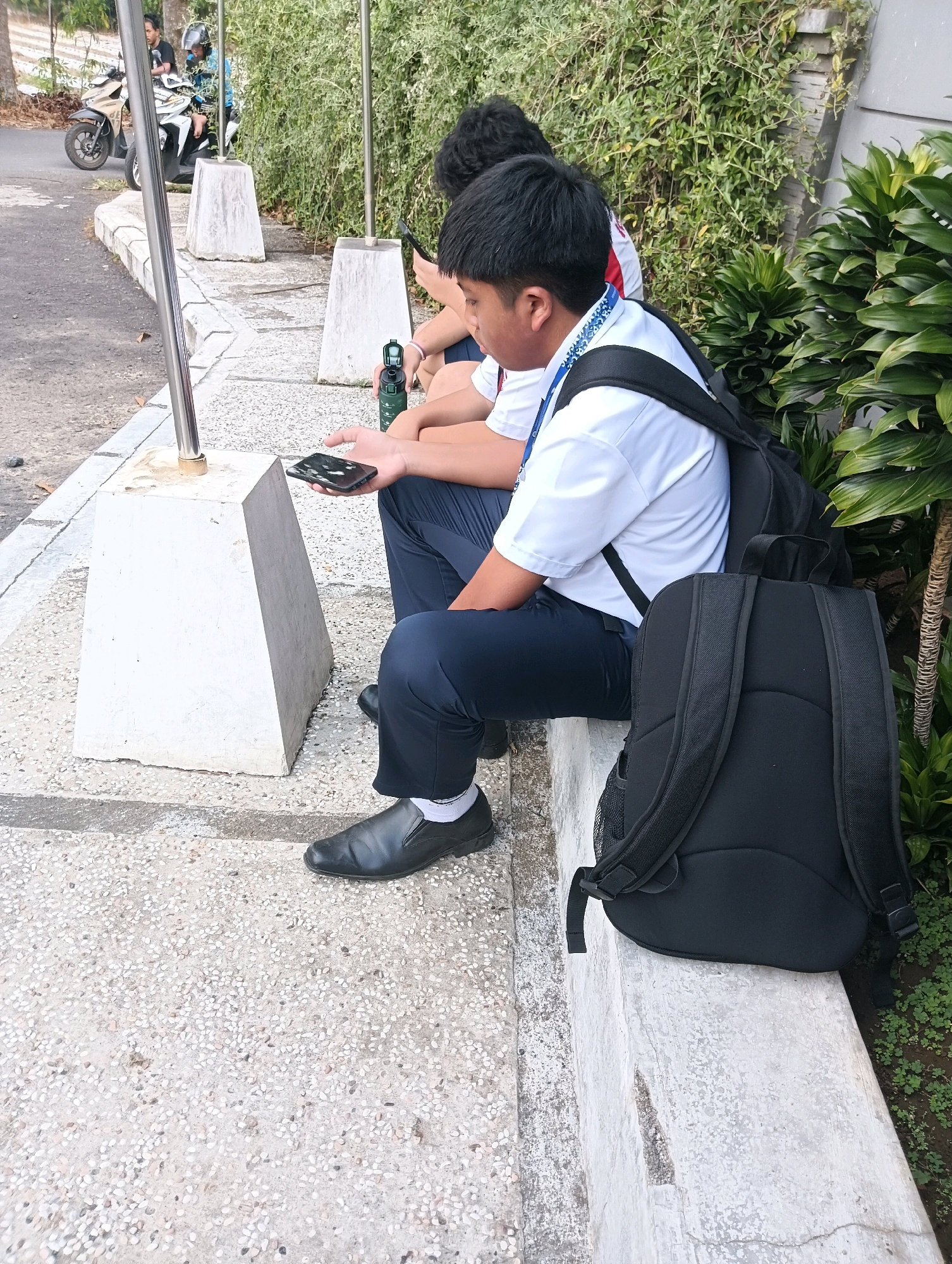 In Tomohon, Indonesia, two young individuals are seated on a concrete ledge outdoors, engaged with their mobile phones. The person in the foreground, identified as male by his attire and hairstyle, wears a white collared shirt, dark blue trousers, black slip-on shoes, and white socks. He has a black backpack on his lap and is holding a dark-colored smartphone. Next to him, another individual, also dressed in a white collared shirt and dark trousers, is partially visible, also holding a phone.
The environment suggests a school or public area, with a paved pathway and a concrete barrier. Green foliage, including leafy plants and trees, borders the seating area, indicating a tropical or sub-tropical setting. In the background, a motorcycle is parked near a pole, and a second, larger tree is visible. The lighting indicates daylight, and the overall scene conveys a casual moment of rest and digital engagement.