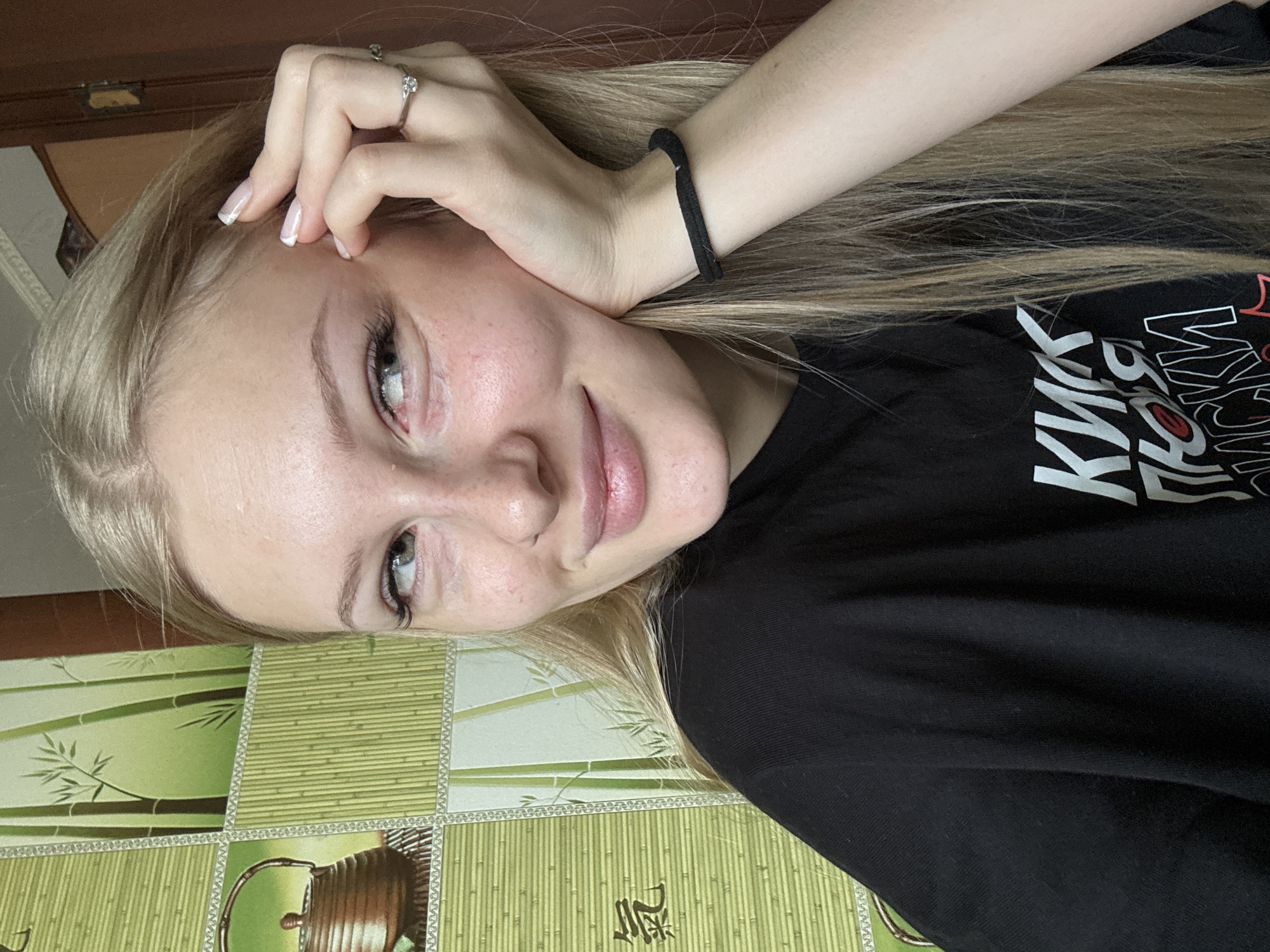 The image shows a young woman with blonde hair, looking to her right with a slight smile. She is wearing a black t-shirt. Her right hand is resting on her cheek, and she has a ring on her index finger and a black hair tie on her wrist. The background features a wall with a pattern of bamboo stalks and what appears to be a decorative teapot. The lighting suggests an indoor environment. There is no information about the location in Комарихинский, Russia.