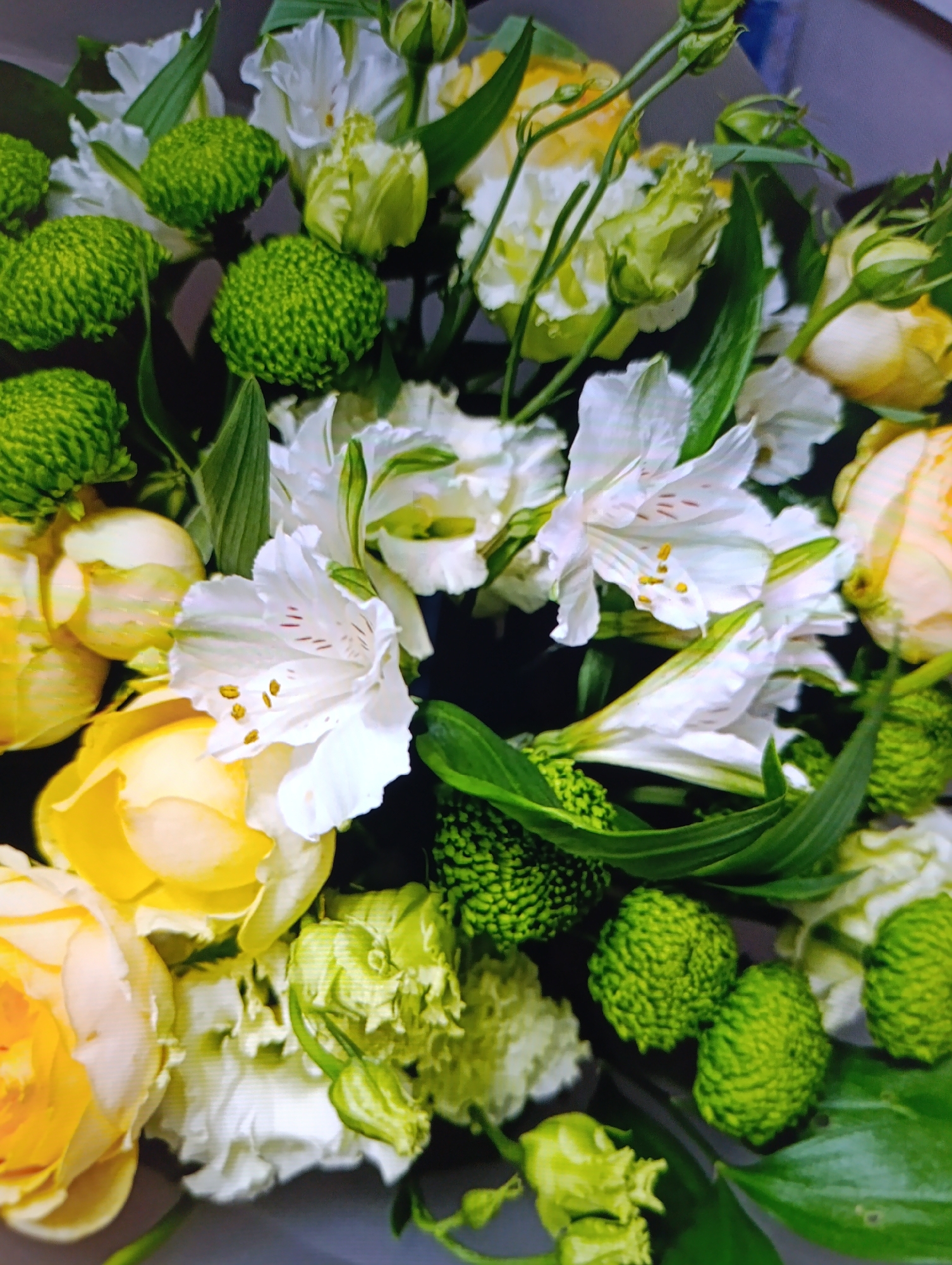 A close-up shot of a floral arrangement. It consists of white Alstroemeria flowers with small orange/yellow markings on their petals, yellow and white roses, and green chrysanthemum pompon flowers. Some unopened buds and green foliage are visible throughout the arrangement. The background is blurred. Location: Зипуново, Russia (location provided by user).