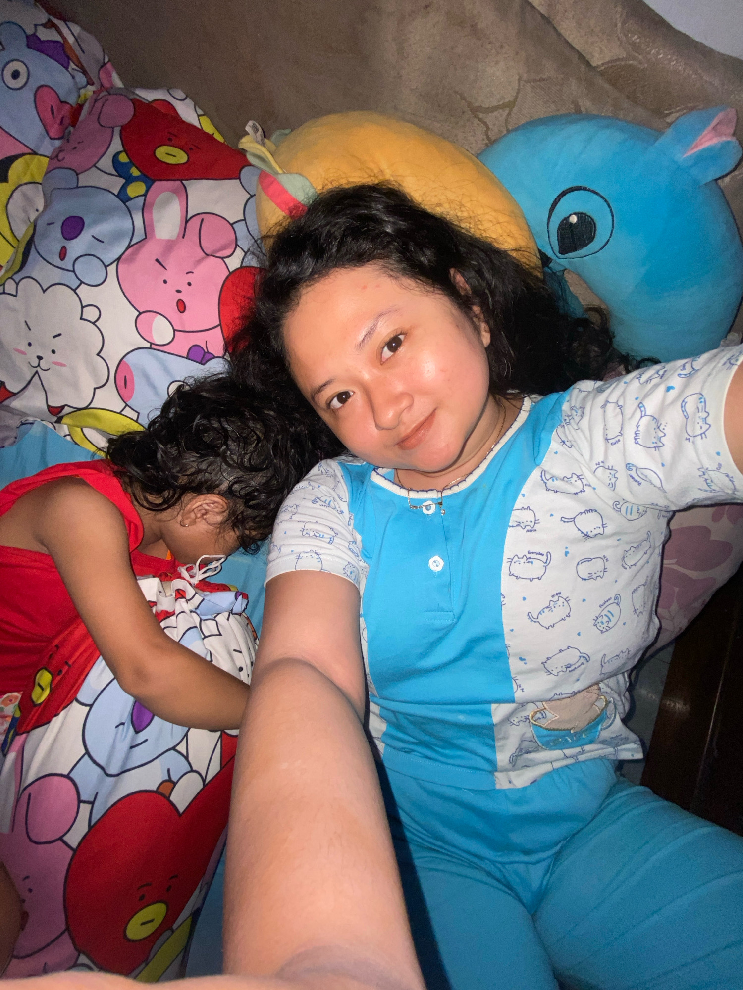 A selfie is taken from a high angle, looking down at two individuals lying on a bed. On the left, a child in a red shirt is asleep, curled up on their side with their head resting on their arm. The bed is covered with a vibrant comforter featuring cartoon characters, possibly from the BT21 franchise. To the right of the child, an adult lies on their back, looking directly at the camera with a slight smile. The adult is wearing a blue and white pajama set adorned with cartoon cats. Behind the individuals are large stuffed animals, a yellow crescent-shaped one and a blue character with large eyes, possibly a Stitch plush. The lighting suggests the room is dimly lit, indicative of nighttime or a curtained room. The overall scene conveys a sense of comfort and domesticity. The image location is stated to be in Gelam, Indonesia.