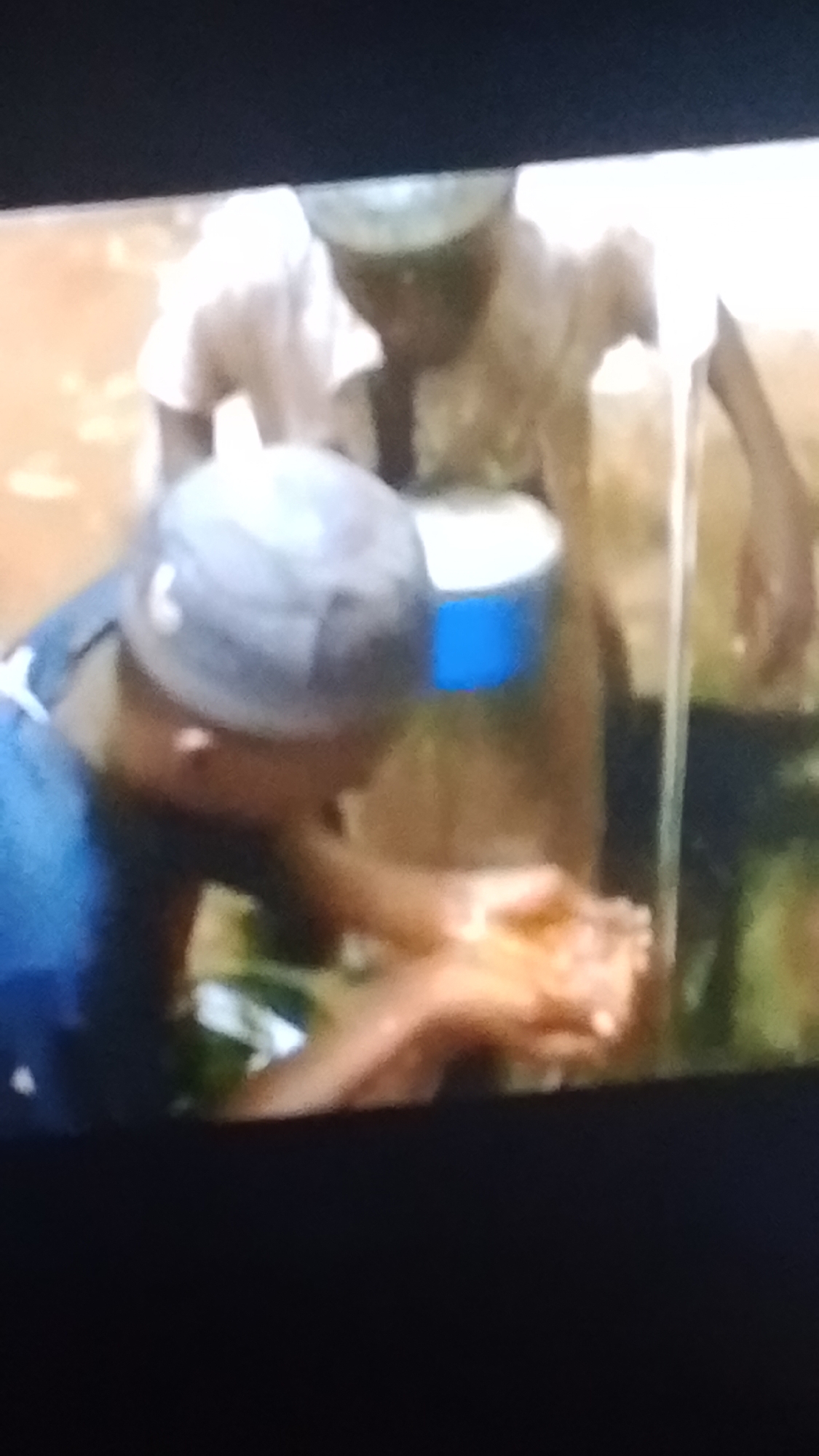 The image shows two individuals, one wearing a blue shirt and a grey cap, the other in a light-colored shirt. The person in the blue shirt is in the foreground, leaning down and washing their hands in what appears to be a natural setting. Their hands are cupped and catching water, suggesting a source of water nearby, possibly from a tap or a natural stream. Behind them, the second person is partially visible, holding a blue and white cylindrical container. The background is blurred but suggests an outdoor environment with some vegetation. The overall scene depicts a moment of handwashing, likely in an outdoor or semi-outdoor area. The image does not contain any specific visual cues that would definitively place it in Lokoja, Nigeria.