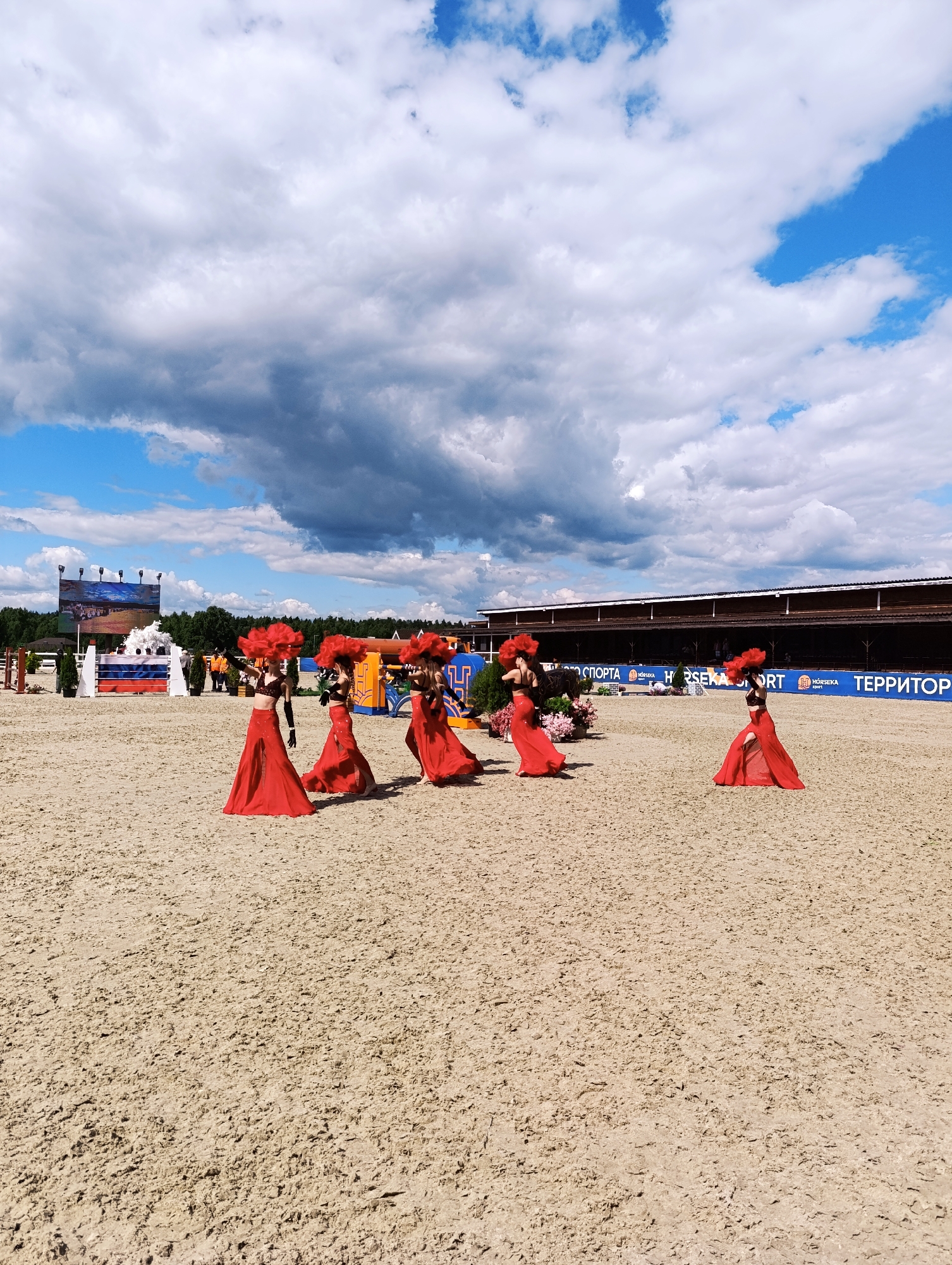 This image captures a performance at an equestrian event in Ivanovskoe, Russia. Five women dressed in elaborate red costumes with large, feathery headpieces are performing on a sand-covered arena. Their costumes consist of red, floor-length skirts with high slits and black, bejeweled tops. They are spread across the arena, with some facing the camera and others looking away. In the background, a large screen displays an image, and beyond that, a forest line is visible. A long, wooden structure, likely a stable or grandstand, runs along the back of the arena. The sky is partly cloudy, with a mix of white and dark grey clouds. The ground is a light brown, finely granulated sand. The overall scene suggests a festive or competitive atmosphere at an outdoor venue.