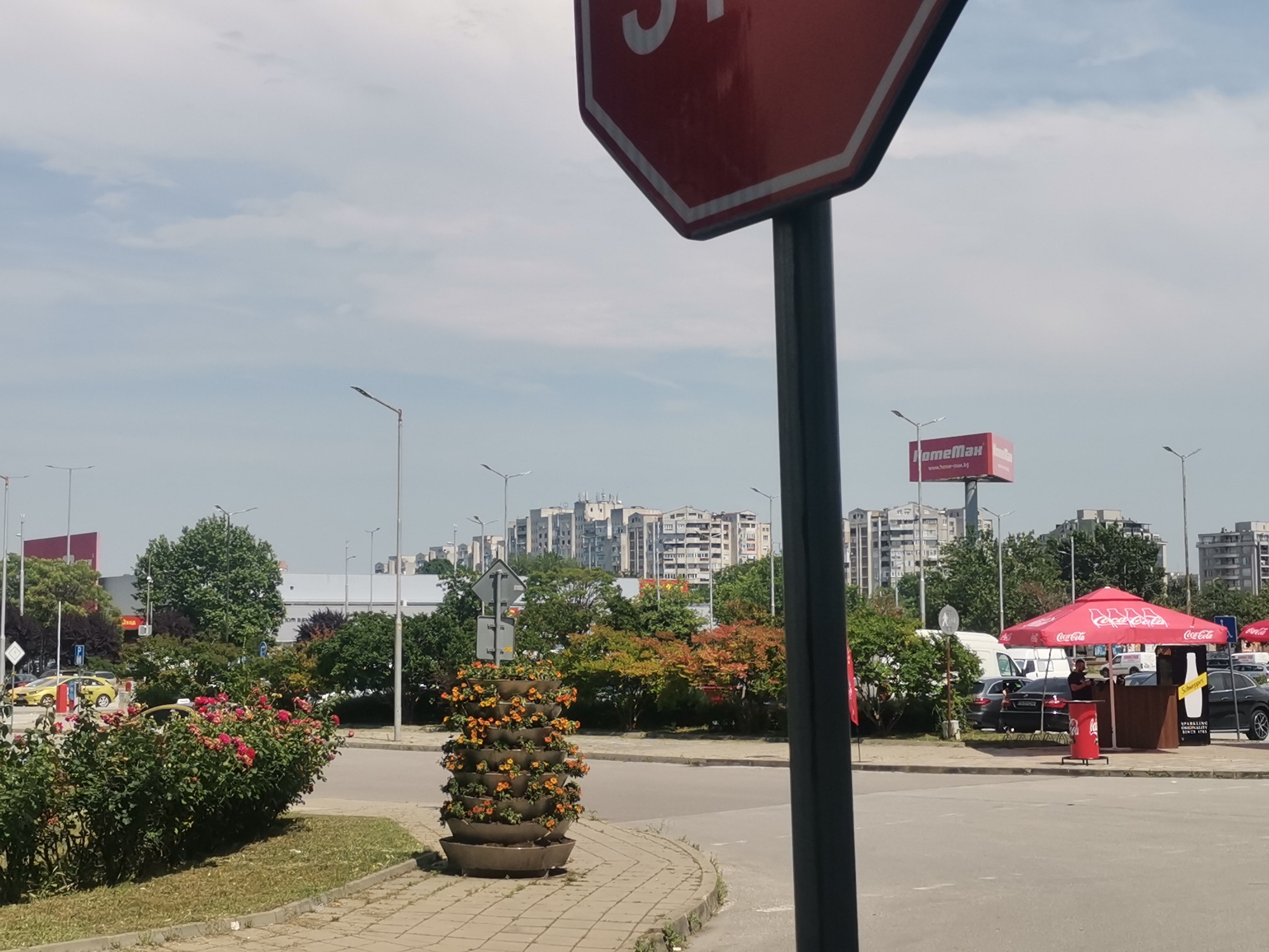 This image depicts an urban street scene in Plovdiv, Bulgaria, specifically near HomeMax at bul. "Osvobozhdenie" 18. The sky is a light blue with scattered, wispy white clouds.
In the immediate foreground on the right, a significant portion of a red octagonal stop sign with white lettering (showing "ST") and its dark pole is visible. To the left, a grassy patch borders a paved sidewalk. On this sidewalk, a multi-tiered concrete planter filled with small orange flowers stands prominently. Adjacent to the planter and grass is a dense bed of rose bushes featuring numerous vibrant pink/red blooms.
The midground is dominated by an asphalt roadway. On the far left, two yellow taxi cabs are partially visible. Further into the midground, beyond the rose bushes and road, are various trees with both green and reddish-orange foliage. On the right side of the road, a small brown kiosk is situated, covered by a large red umbrella branded with multiple "Coca-Cola" logos. An adult male is standing behind the counter of this kiosk, facing left. A red Coca-Cola receptacle is positioned next to the kiosk. Several vehicles, including a dark sedan and a white van, are parked near the kiosk.
In the background, a large billboard with the "HomeMax" logo and "www.home-max.bg" is visible above the trees and a building structure. Further left, another red commercial sign can be seen. The distant background features numerous multi-story residential apartment buildings with light-colored facades. Multiple streetlights with single luminaires are scattered throughout the scene.
