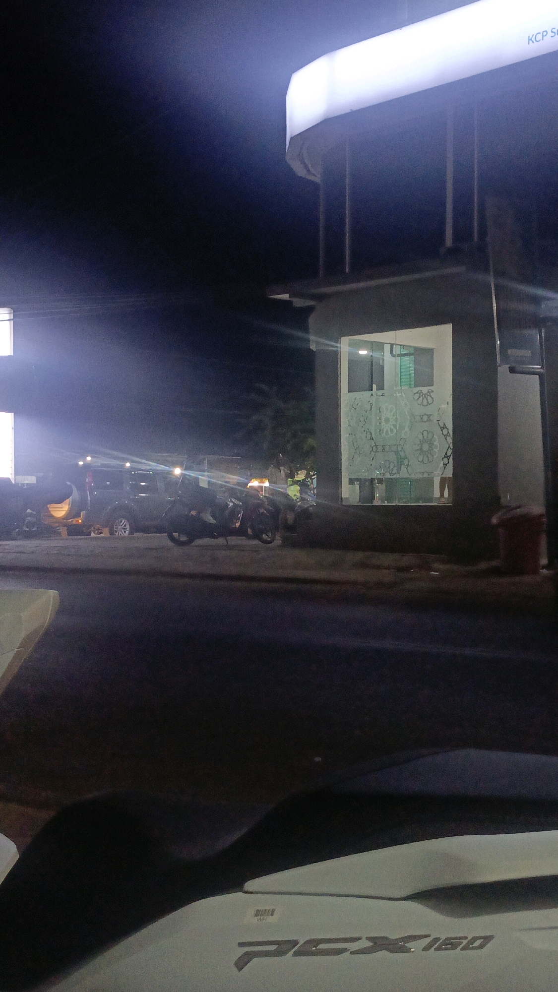 This nighttime photograph, taken in Taliwang, Indonesia, captures a dimly lit street scene. In the foreground, a portion of a white scooter with the visible text "PCX160" is present. The mid-ground shows a street with parked vehicles including a dark-colored SUV and two motorcycles. The background features a building with a bright, illuminated sign reading "KCP Su". The building also has a glass-paneled entrance with decorative patterns. A red trash bin is visible next to the building. The overall ambiance is that of a quiet urban environment at night.