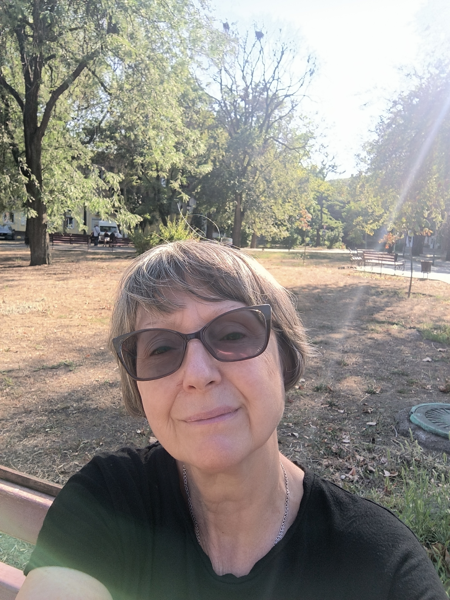 A middle-aged woman wearing sunglasses and a black shirt is taking a selfie in a park. She has short, graying hair and is smiling. The park appears to be in a city, with trees, benches, and a path visible in the background. The sun is shining, casting a warm glow on the scene. The image also shows a manhole cover on the ground. The location is the city of Mykolaiv, Ukraine.