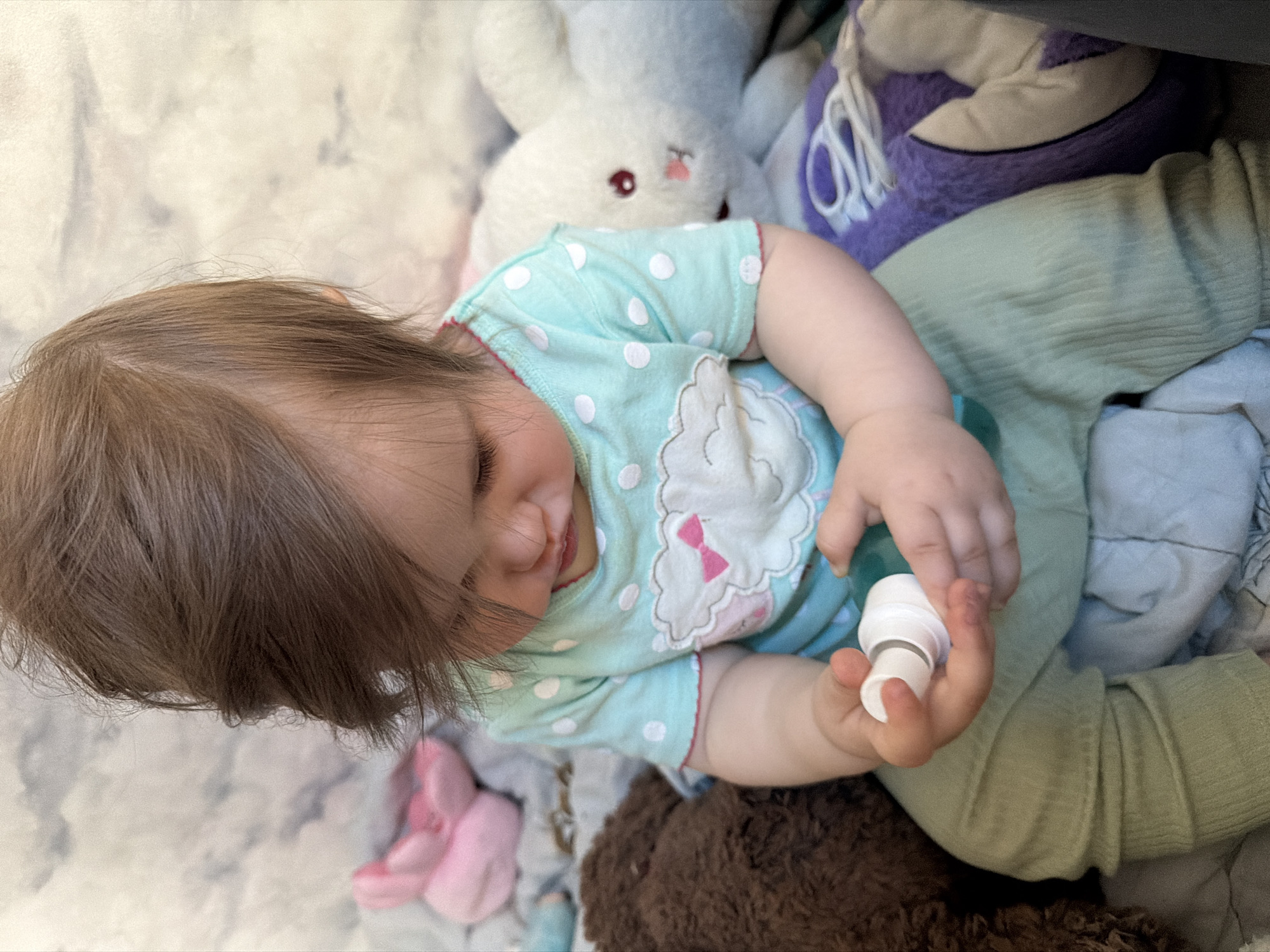 A child lies on a patterned surface surrounded by stuffed animals. The child is wearing a light blue, polka-dotted onesie with a cloud design on the chest and light green pants. They are holding a small, white object in their right hand. The stuffed animals include a white bunny, a purple and white plush object, and a brown teddy bear. The location is unspecified and not identifiable as Tokkari-Lend, Russia.