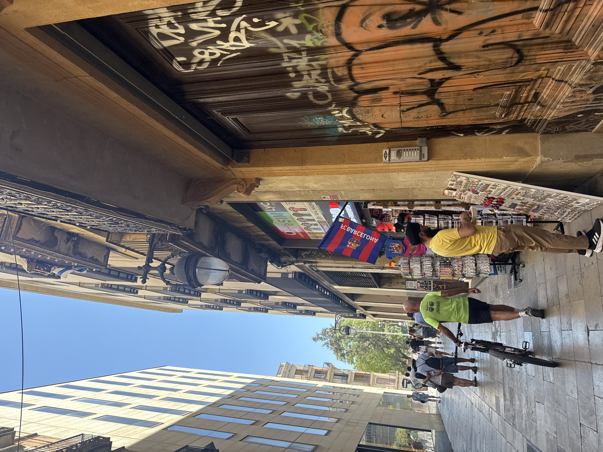 The image depicts a sunlit pedestrian street scene in Barcelona, Spain. On the left, a modern building with rows of rectangular windows stands tall, contrasting with the older, more ornate architecture on the right. The building on the right features balconies with intricate iron railings and displays a shop with a colorful Catalan flag hanging outside. Graffiti adorns the large wooden double doors of a building on the right side of the street. Several people are walking along the cobblestone path. In the foreground, a man wearing a bright green shirt and black shorts pushes a bicycle, while another man in a yellow shirt stands near a stall displaying various merchandise. The overall atmosphere is that of a bustling urban environment on a clear day.