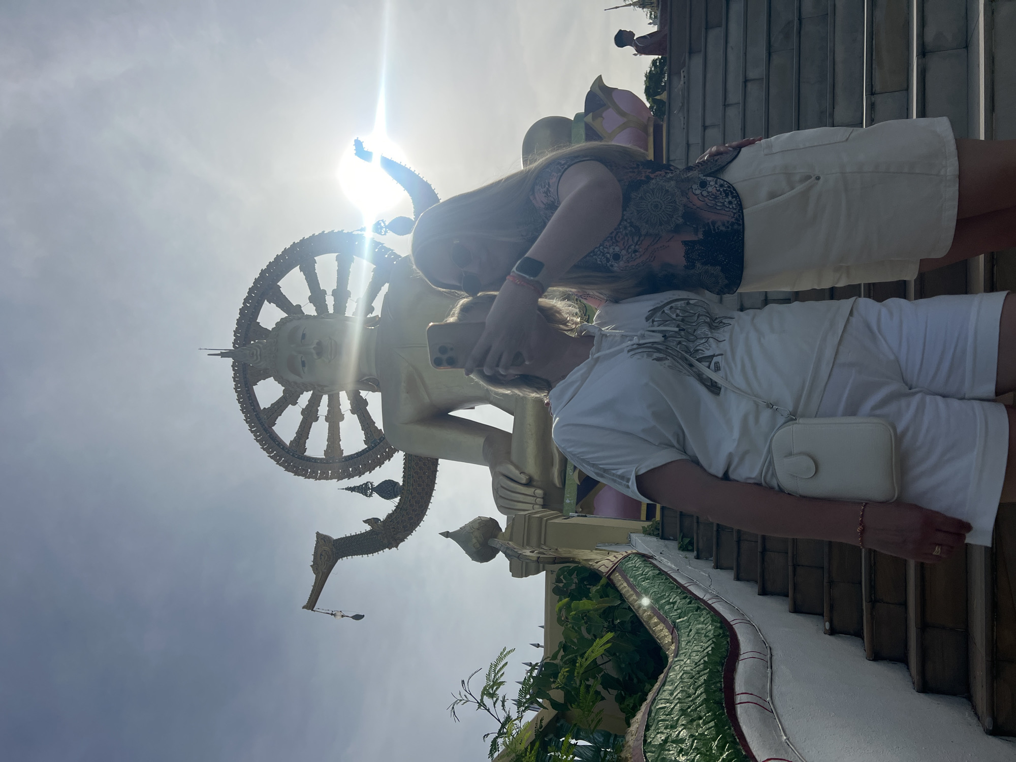 Two women are standing on stairs in front of a large golden Buddha statue in Baan Plai Laem, Thailand. The woman on the left is wearing a white t-shirt and white shorts, and has a white crossbody bag. The woman on the right is wearing a patterned top and white shorts, and sunglasses. They are taking a selfie with a smartphone. The sky is bright and sunny, with some lens flare from the sun. The Buddha statue is very large and imposing, with a wheel of dharma behind its head. The stairs are decorated with green serpent statues on either side. There are other people in the background, including a child.