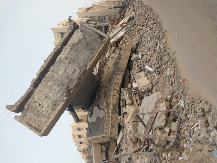 The image displays a street scene in Khan Yunis, Palestinian Territory, characterized by extensive structural destruction under a clear, pale sky. In the foreground and midground, a substantial pile of grey concrete rubble, fragmented building materials, and earth dominates the left side. A large, fractured reinforced concrete slab, revealing exposed rebar, is tilted steeply upwards from this debris pile, pointing towards the upper left. Scattered smaller debris, including plastic fragments, are visible within the rubble and along its base. To the right, a light brown paved road curves along the edge of the debris. In the background, a row of multi-story, light-colored residential buildings stands, appearing largely structurally intact, with visible windows and balconies. No individuals are present in the scene.