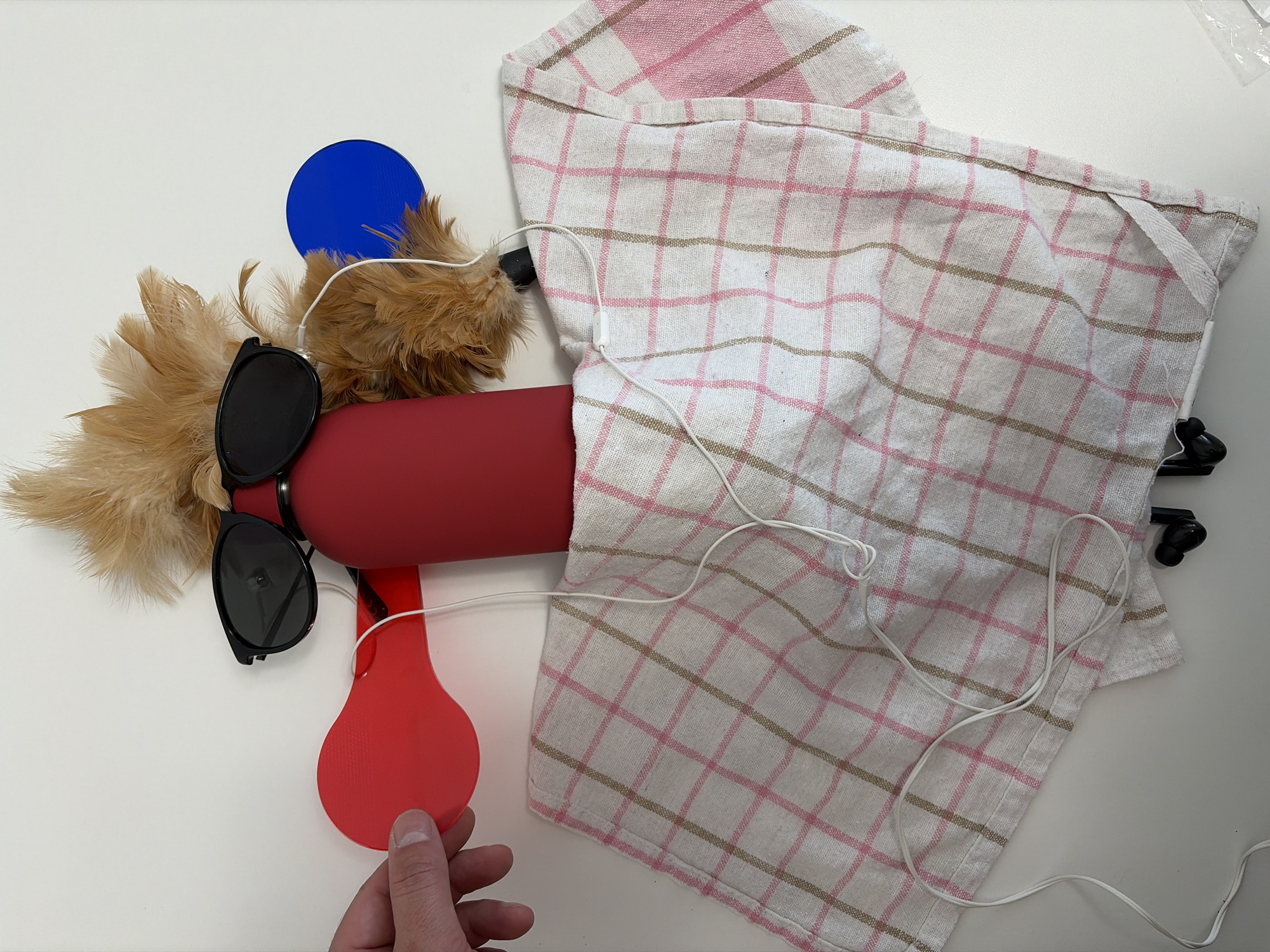 The image presents an artistic or whimsical arrangement of everyday objects on a clean, white surface, presumably indoors in Budva, Montenegro. Central to the composition is a red, cylindrical object, resembling a bottle or a dispenser, lying horizontally. A pair of black sunglasses is perched on top of this red object, facing left, giving it a humanoid or creature-like appearance.
Attached to the left end of the red cylinder, behind the sunglasses, is a cluster of soft, light brown feathers, similar to those found on a feather duster. To the upper left of these feathers, a small, circular blue object stands upright.
A white and pink plaid dishtowel or tea towel is draped over the right side of the red cylinder and covers a large portion of the right side of the frame. A thin, white cable, possibly from headphones or a charging device, is casually laid across the towel, extending towards the feathery end of the red object.
In the bottom left corner, a person's hand, with neatly kept fingernails, is seen holding a red, paddle-shaped object, possibly a small spatula or part of a toy. This interaction suggests that the objects are being arranged, played with, or are part of a creative setup.
The lighting is bright and even, indicating a daytime indoor setting. No specific weather conditions are discernible, and no text is visible within the image. The overall scene conveys a sense of playful assembly or an abstract representation using common household items.