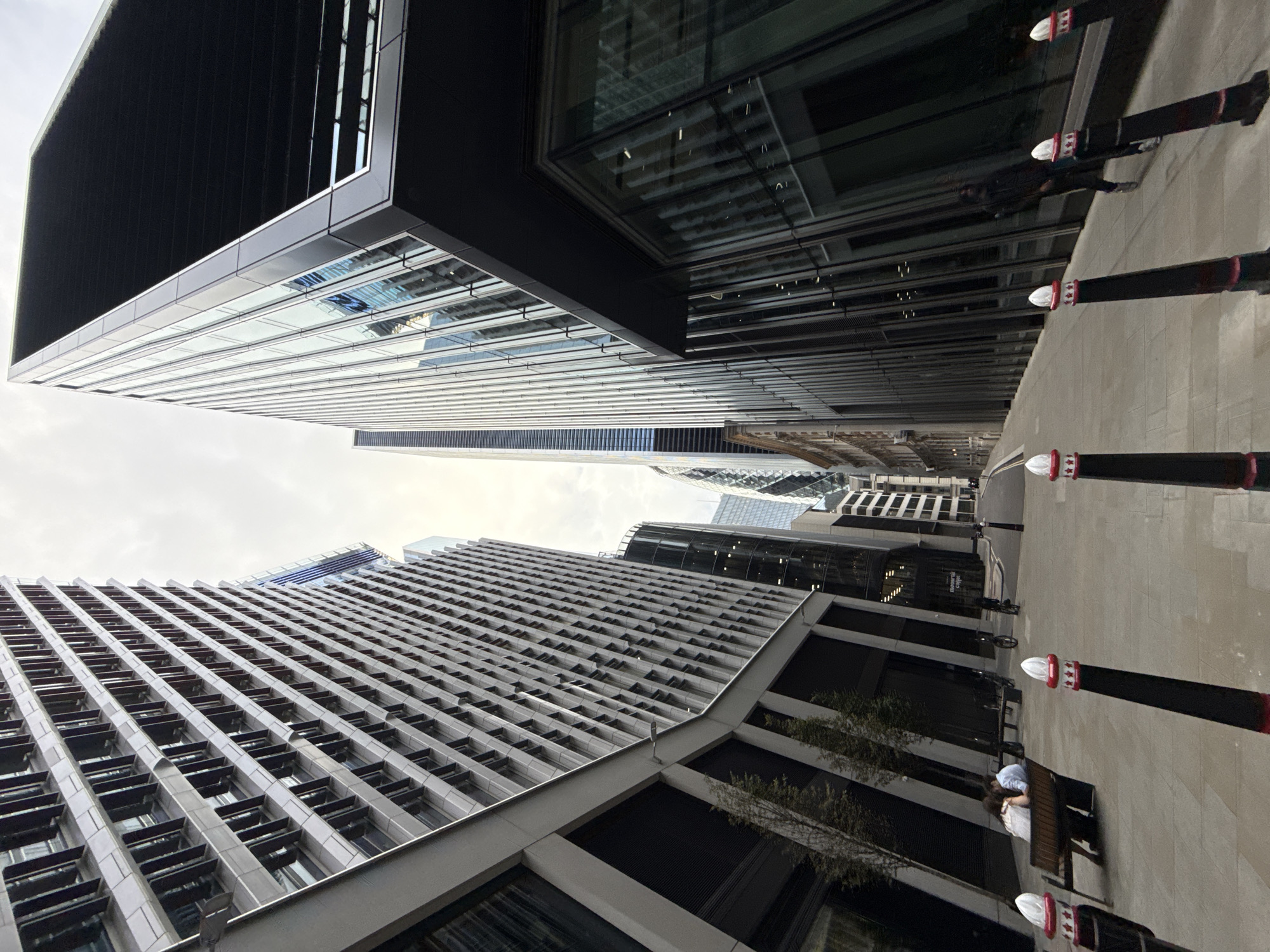 The image depicts a narrow street in the City of London, United Kingdom, flanked by modern skyscrapers. On the left, a tall building with a grey, multi-paneled facade dominates the view, its angled structure creating a sense of depth. To the right, a striking, dark-colored skyscraper with a distinctive angled roof and reflective glass facade stands prominently. Further down the street, other modern buildings with glass and steel exteriors are visible, including the iconic Gherkin building.
In the foreground, a paved pedestrian walkway is lined with decorative bollards. A wooden bench is placed along the left side of the walkway, occupied by two individuals sitting close together. A bicycle is parked nearby. Further down the street, another person is seen walking, and a bicycle is ridden by someone. Trees are planted along the left side of the walkway, interspersed between the buildings. The sky is overcast, suggesting a cloudy day. The architecture and urban planning suggest a financial district.