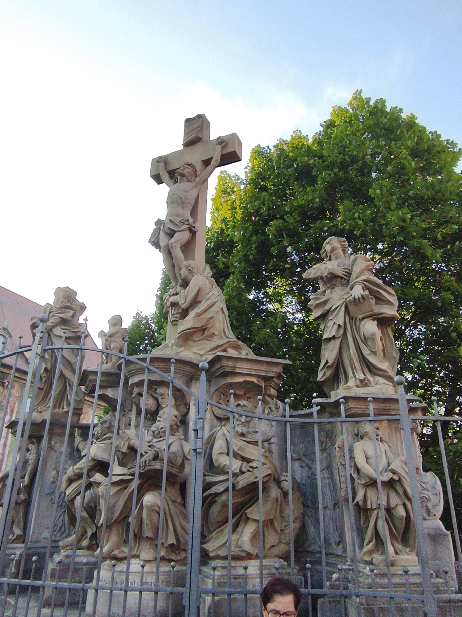 The image depicts a Christian monument, specifically a crucifixion scene, located in Bamberg, Germany. A large stone crucifix, with Jesus Christ depicted, stands prominently at the top. Surrounding the crucifix are several other stone statues of figures in religious attire, some seated and others standing. These statues appear to be part of a larger sculptural ensemble, possibly an altar or a memorial. A wrought-iron fence encloses the monument, with some of the statues visible behind it. In the background, lush green trees provide a natural backdrop, and a building with a red-tiled roof is partially visible to the left. The sky is bright and clear, suggesting daytime. The overall scene conveys a sense of historical and religious significance.