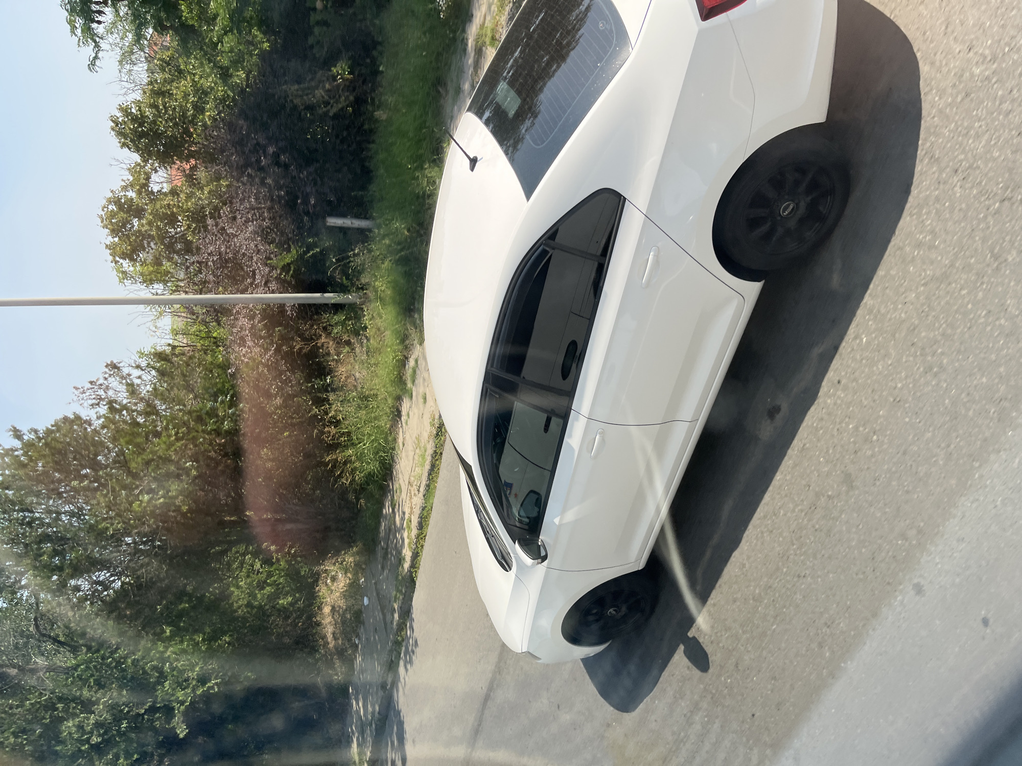 A white sedan is parked on a paved road next to a patch of dry grass and trees. The car's windows are tinted, and it has black rims on its wheels. The scene appears to be in Agigea, Romania, given the context. The sun is shining, casting shadows on the road. The background features lush green trees and shrubs, with a clear blue sky overhead.