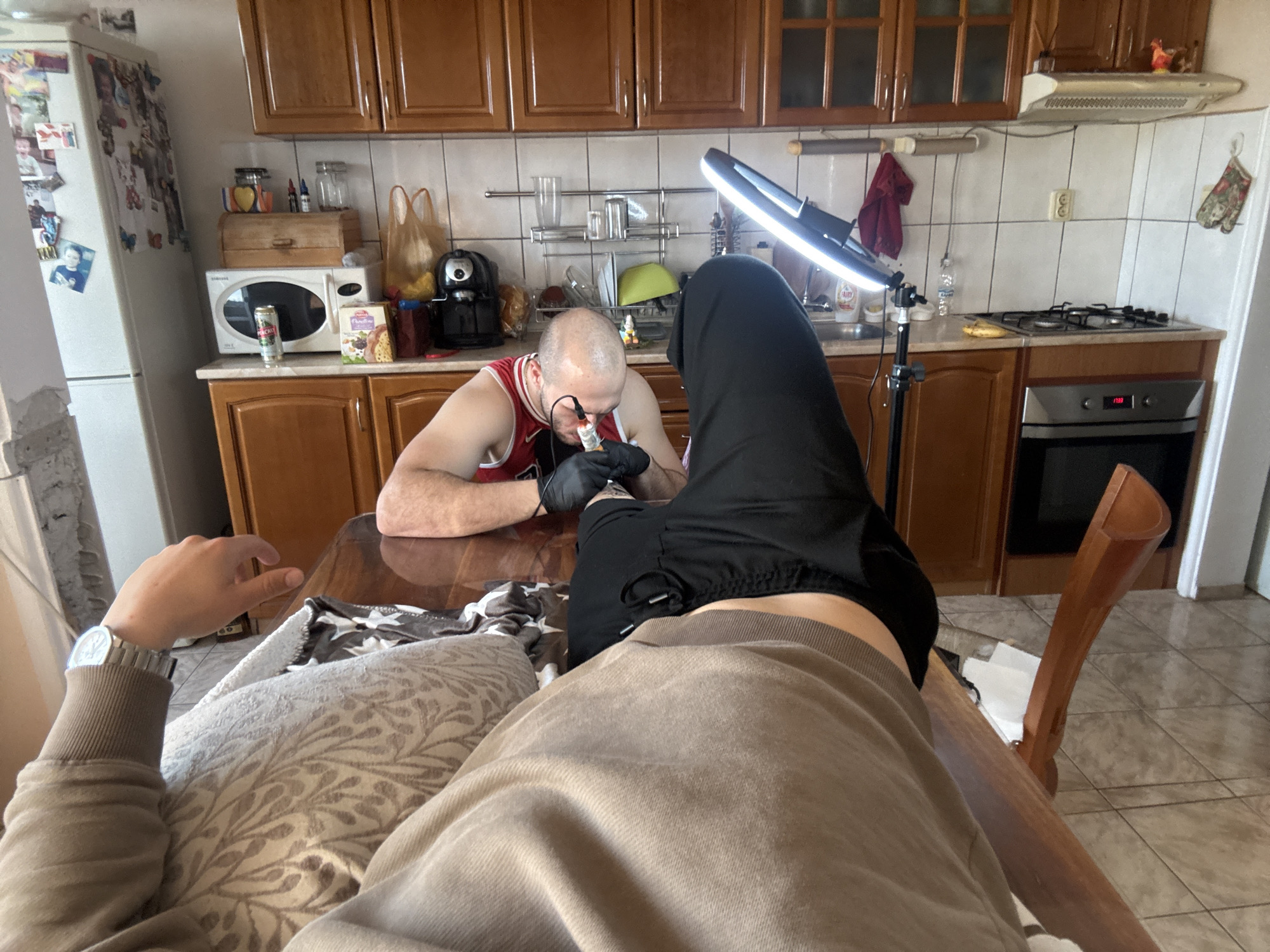 The photograph shows an indoor scene inside a kitchen. A person is lying on a table, with only the upper part of their body and legs visible. They are wearing a light brown sweatshirt and dark sweatpants. The photographer's perspective is from the person lying on the table.
A man is seated at the table, leaning forward and performing a tattoo on the person's foot. He is wearing a red and white striped tank top and glasses. He has black gloves on and appears to be focused on his work.
A ring light on a stand is positioned to illuminate the tattooing area. Various kitchen appliances and items are visible in the background, including a refrigerator, microwave, cabinets, stove, and other typical kitchen items. The kitchen has tile floors and a white tiled backsplash.
There is nothing visible in the scene to indicate a specific location in the city of Burgas, Bulgaria.