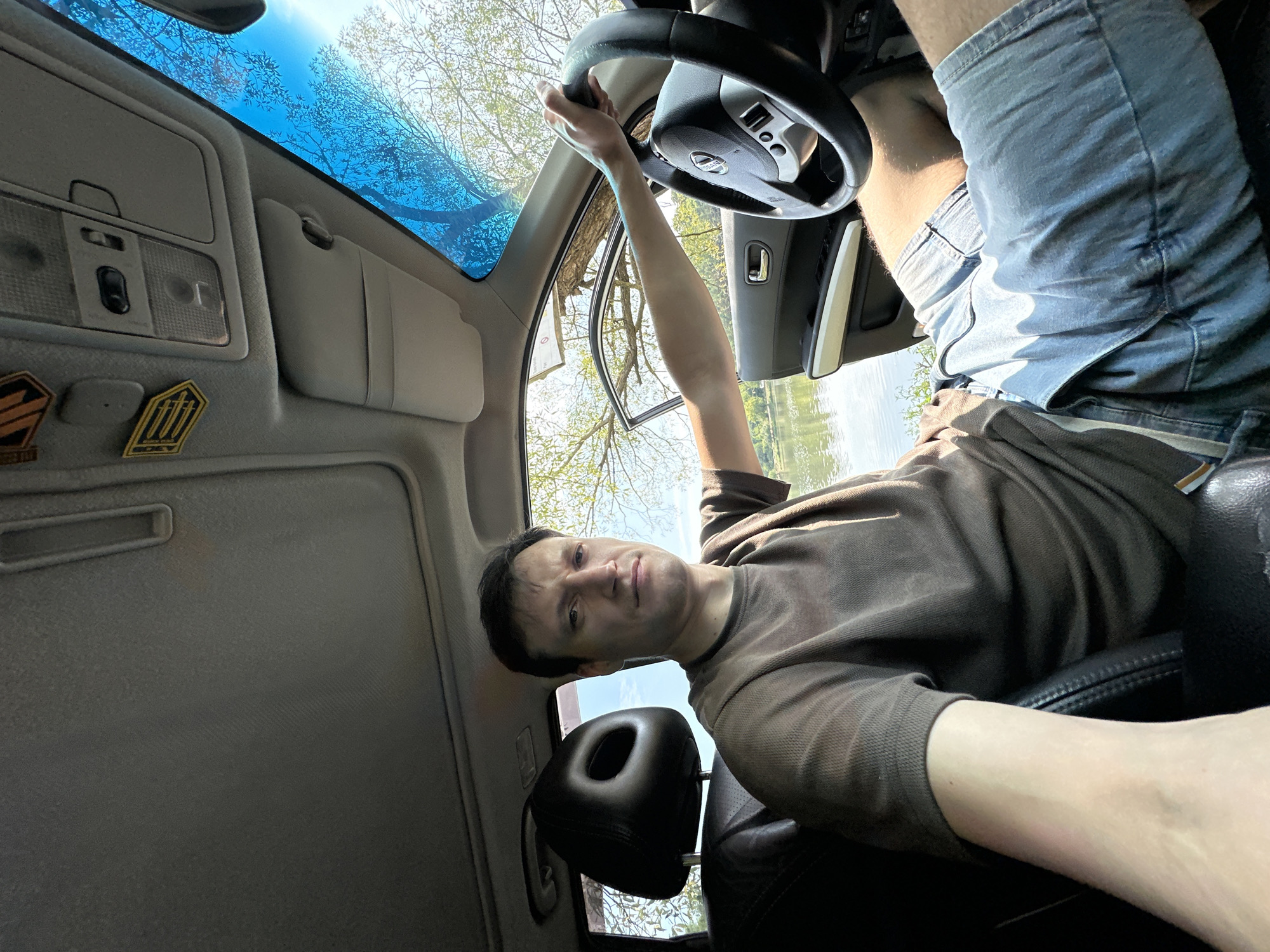 A man is seated in the driver's seat of a car, which is parked outdoors near a body of water. The car's interior is visible, including the steering wheel, dashboard, and a sunroof. The man is wearing a brown t-shirt and denim shorts. His right arm rests on the steering wheel, and his left arm is extended towards the passenger side of the car. The car door on the passenger side is open, revealing trees and a body of water outside. The sky is blue with some clouds. The man is looking directly at the camera with a neutral expression. The location is identified as Makiv, Ukraine.