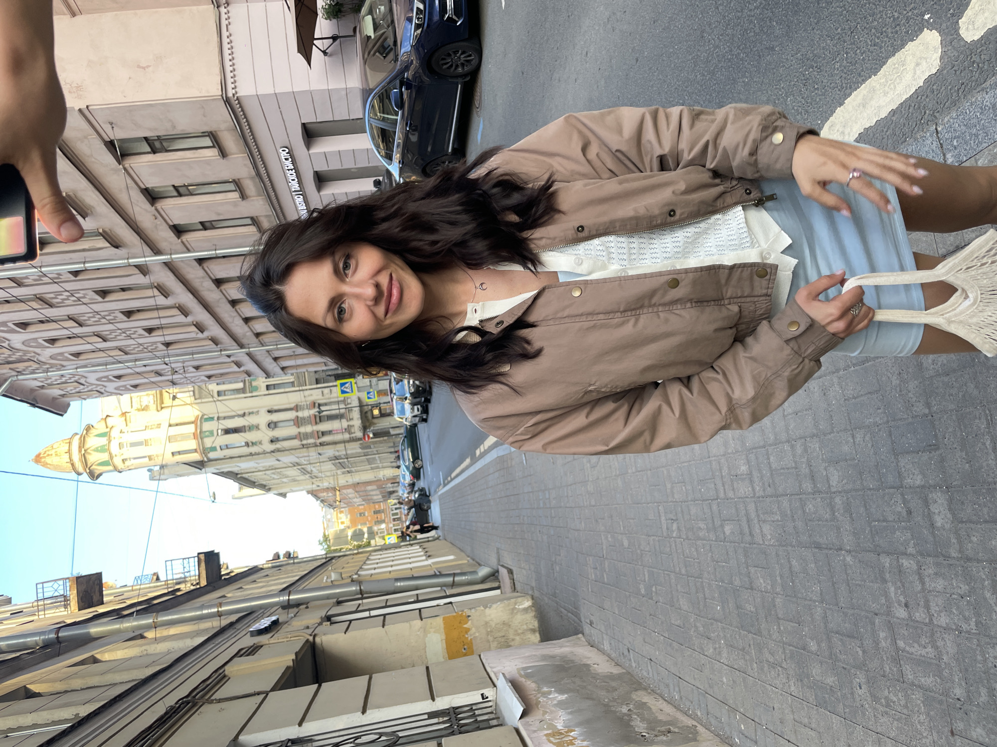 A young woman with dark hair, wearing a light blue skirt, a white textured top, and a tan bomber jacket, stands on a cobblestone sidewalk in Saint Petersburg, Russia. She is holding a white mesh bag in her left hand and has a ring with a pink stone on her right ring finger. In the background, cars are parked along a street lined with multi-story buildings. One building features intricate architectural details and a conical tower. The sky is clear and blue, indicating daytime.