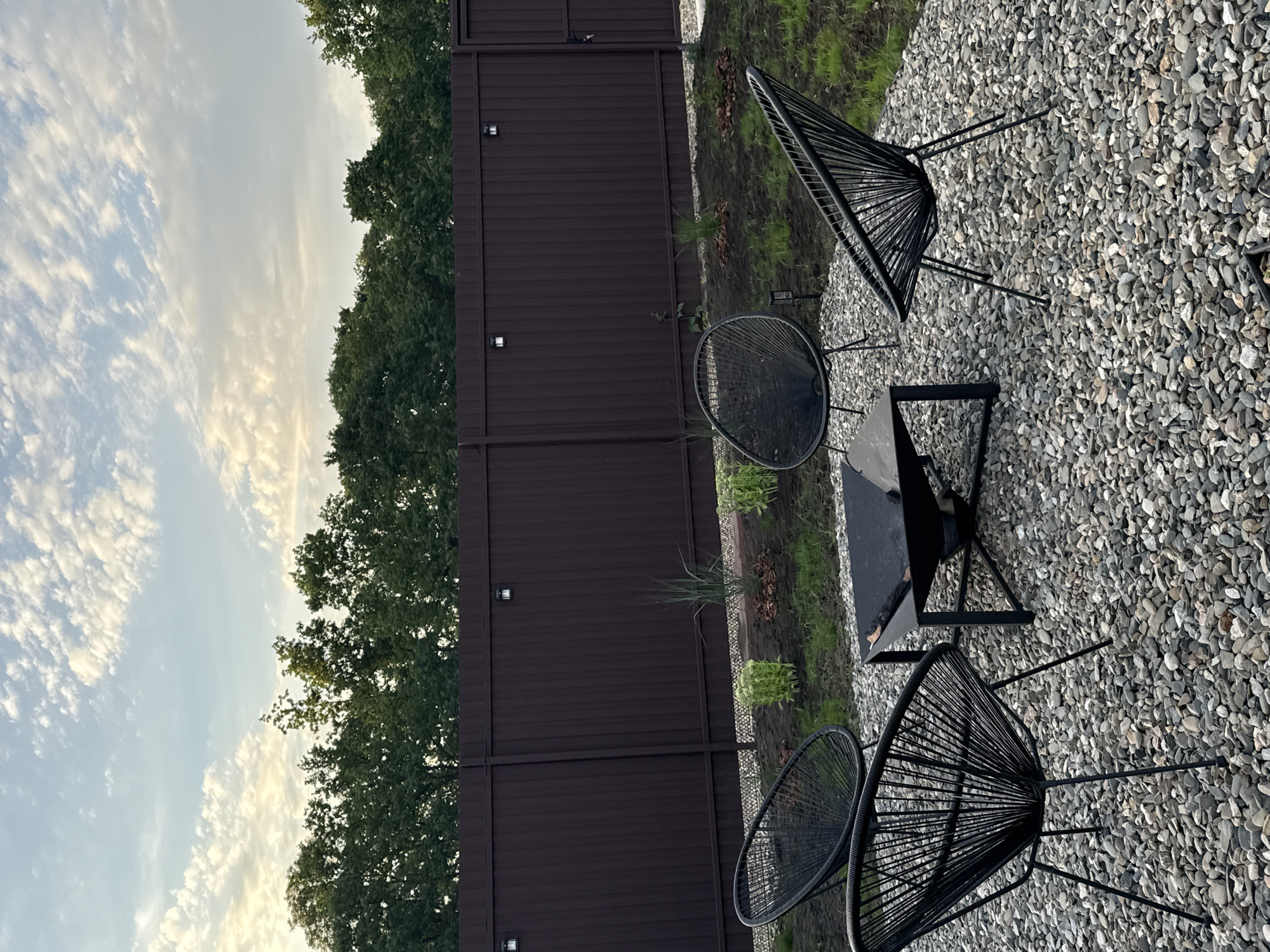 In the city of Starokorsunskaya, Russia, a modern outdoor seating area is depicted. The scene is set on a gravel patio, surrounded by a dark brown metal fence. The sky above features scattered white clouds against a pale blue backdrop. Four black wire-frame chairs, in a distinctive Acapulco style, are arranged around a black metal fire pit. The fire pit has a square top and a geometric base. Beyond the seating area, the ground transitions to soil and grass, with small plantings of greenery and mulch. The fence is punctuated by several small, square outdoor lights. The overall impression is of a contemporary, minimalist outdoor living space.