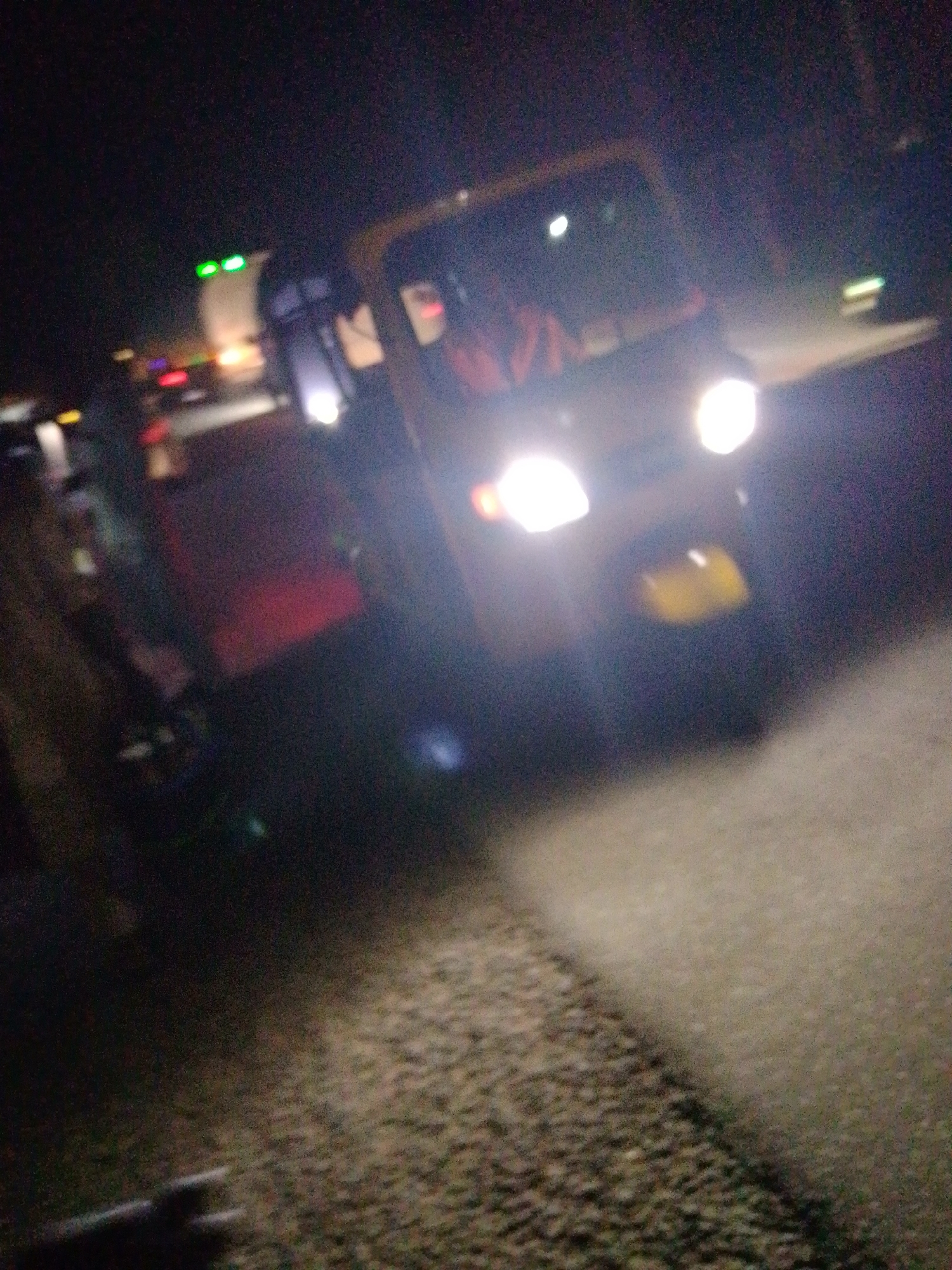 The image is a night-time, blurry photograph of a street scene in Jalingo, Nigeria. A yellow autorickshaw, also known as a "Keke Napep" in Nigeria, is the primary subject, illuminated by its headlights. The driver, a person with a light-skinned complexion and wearing a white shirt, is visible in the driver's seat. The background is dark with other vehicles and lights, suggesting a busy road. The ground appears to be paved with some gravelly sections. The overall impression is of a vehicle moving at night in an urban environment.