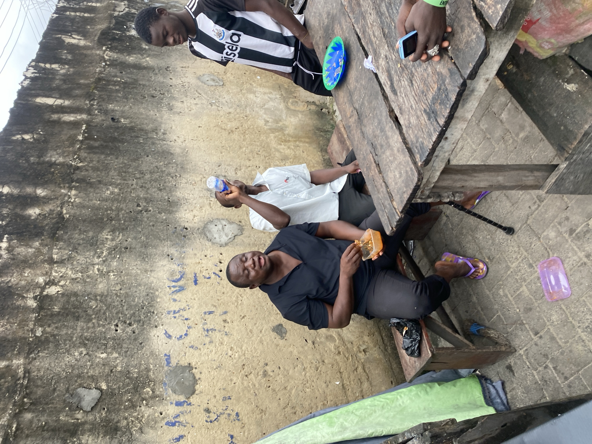 In an outdoor urban setting with a weathered, graffitied concrete wall as the backdrop, three young Black men are gathered around a rustic wooden table. The man on the left, wearing a black polo shirt and black pants, is seated on a wooden bench, eating from an orange plastic container. He has dark skin, a round face, and is looking down at his food. To his right, another man in a white short-sleeved shirt is standing and drinking from a clear plastic water bottle. Further to the right, a third man in a black and white striped football jersey, possibly a Newcastle United kit, stands with his hands in his pockets. A blue and green plastic plate with a white napkin is on the table in front of him. The ground is paved with grey bricks. Various objects are scattered around, including a black plastic bag on the bench, a discarded purple plastic container on the ground, and a light blue mobile phone being held by someone whose hands are visible in the bottom right corner, wearing a green wristband. A green tarp is leaning against the wall to the left of the scene. The overall impression is of a casual, informal gathering in a somewhat dilapidated urban environment.