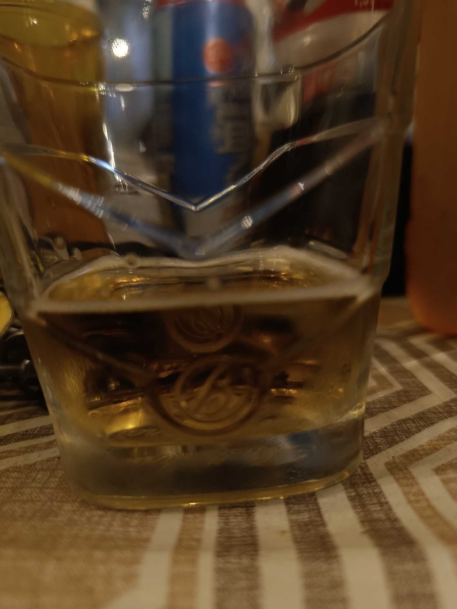 A close-up, eye-level shot captures a partially filled glass of amber liquid, likely beer, resting on a patterned tablecloth. The glass has a distinct angular design around its upper rim and a slight condensation effect on its lower half. Inside the glass, the liquid exhibits frothy foam at the surface and reflects light, revealing embossed patterns on the glass's interior base. In the background, out of focus, are hints of other containers, including a blue and red can, and a blurred orange object. The scene appears to be indoors, possibly a domestic setting or a casual establishment. There is no identifiable information regarding the location within Taraz city administration, Kazakhstan, in this image.