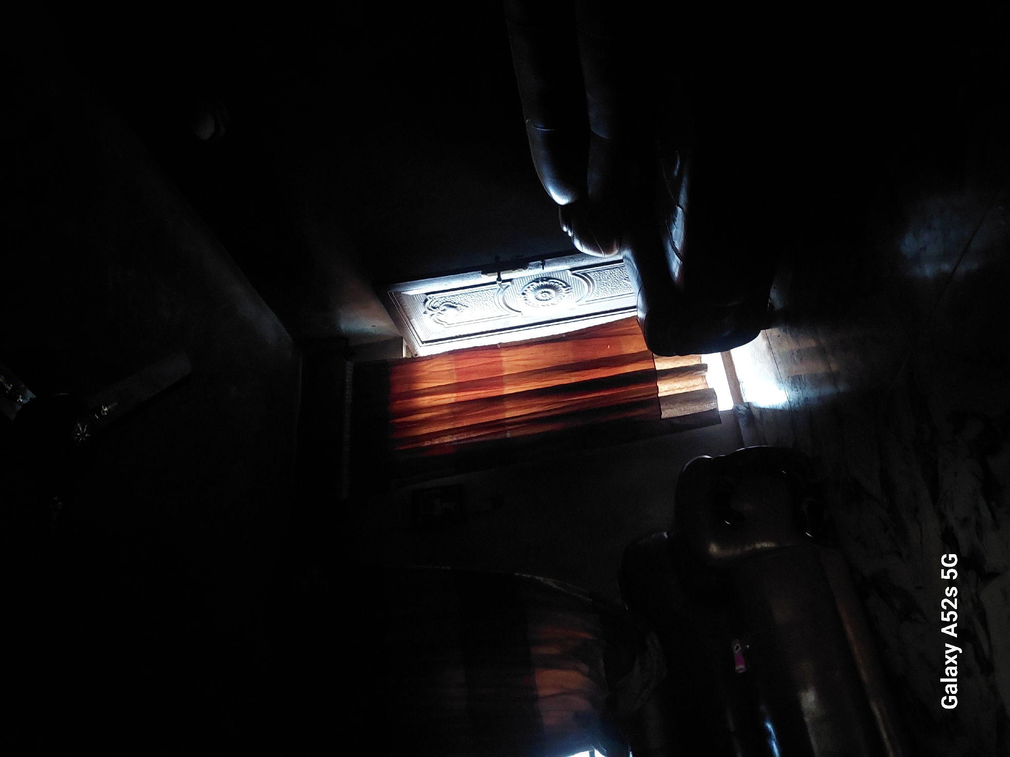 The image shows an interior scene, likely a dimly lit room, with a strong light source coming from above and to the left. The camera is angled such that the scene appears upside down. In the upper portion of the image, a sculpted decorative ceiling or panel is visible, illuminated by the light. Below this, there are horizontal wooden elements, possibly blinds or slats, also catching the light.
To the right, the dark silhouettes of what appear to be legs of a person or large statue are prominent, with distinct shapes suggesting knees and feet. A portion of a shiny, dark object, possibly furniture or another sculpture, occupies the lower right side of the frame. The overall lighting is dramatic, with deep shadows and bright highlights, suggesting either a natural light source like a window or an artificial light positioned to create this effect.
The text "Galaxy A52s 5G" is visible vertically along the right edge of the image, indicating the device used to capture the photo. The context of Awkuzu, Nigeria, suggests a tropical or subtropical climate, but the image itself does not provide direct clues about the weather or time of day beyond the strong light. The scene's ambiance is mysterious and somewhat austere due to the low light and abstract forms. No specific activity is evident; it appears to be a static composition focusing on light and form within a dark interior.